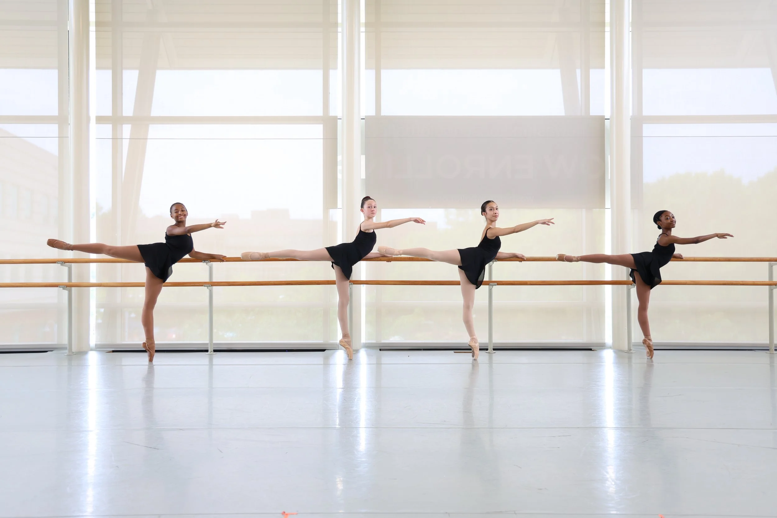 Four ballet dancers in black tutus practicing at a ballet barre inside a dance studio with large windows and natural light.