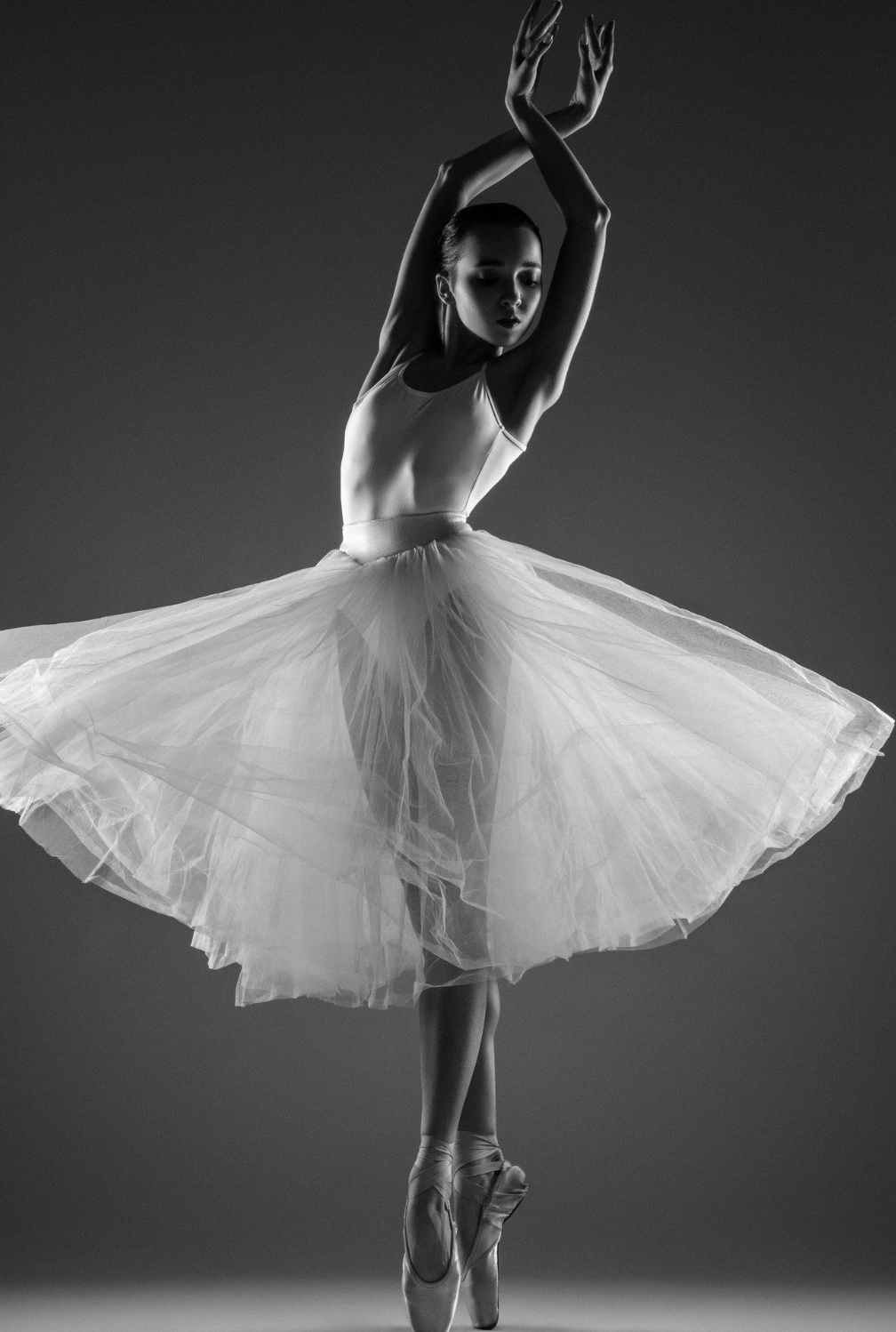 A ballet dancer on pointe wearing a tutu, captured in a graceful pose with arms raised above her head, in black and white.