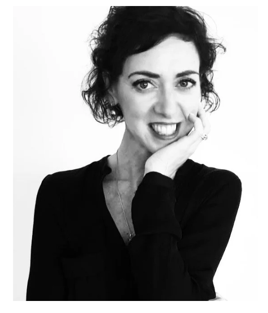 Black and white photo of a woman with short curly hair, smiling and resting her chin on her hand, wearing a dark top.