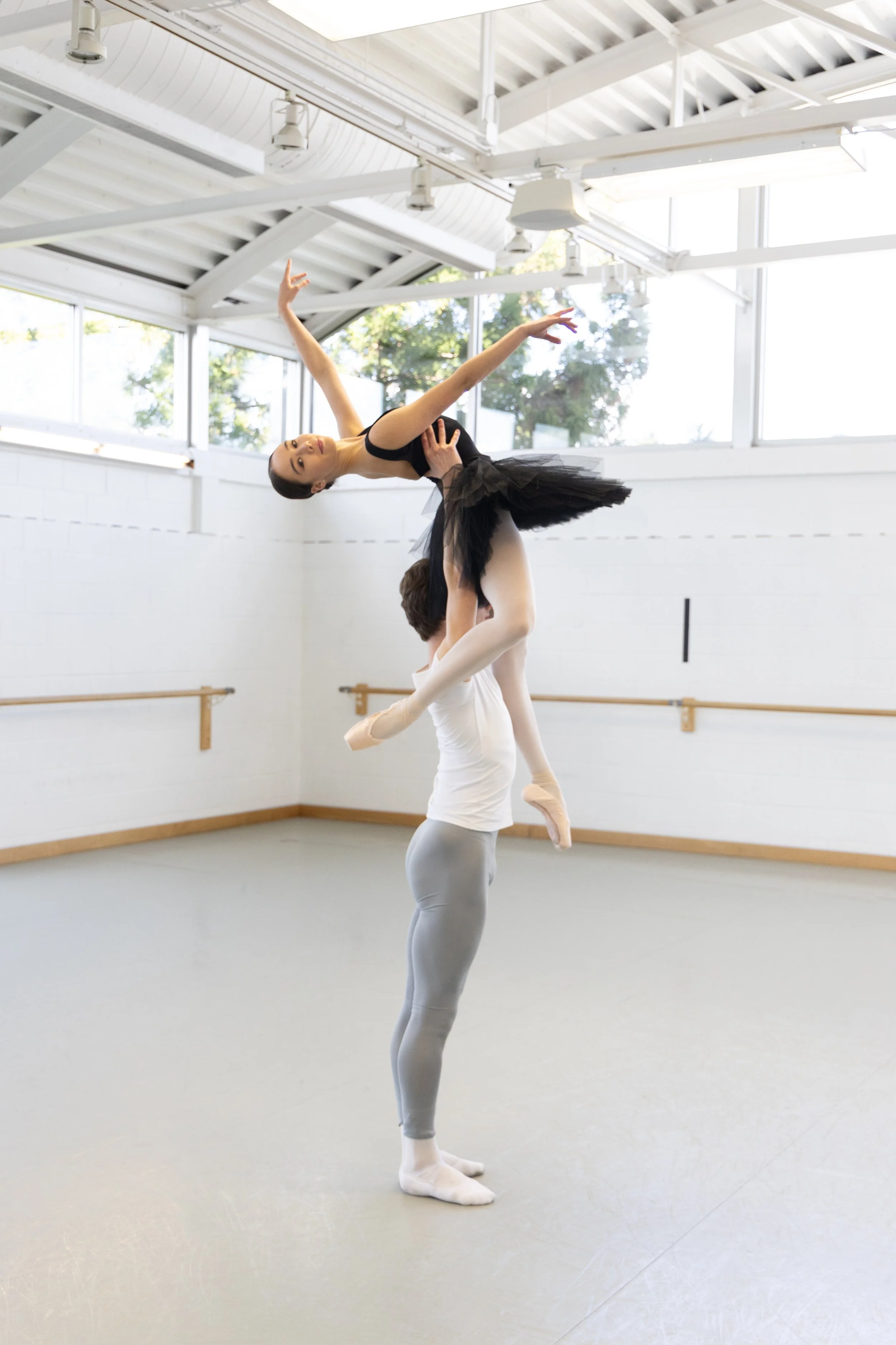 Two ballet dancers practicing in a bright dance studio, one standing on the floor and lifting the other in the air. Both are wearing ballet attire, with the dancer on the floor in gray tights and a white top, and the lifted dancer in a black leotard and tutu.