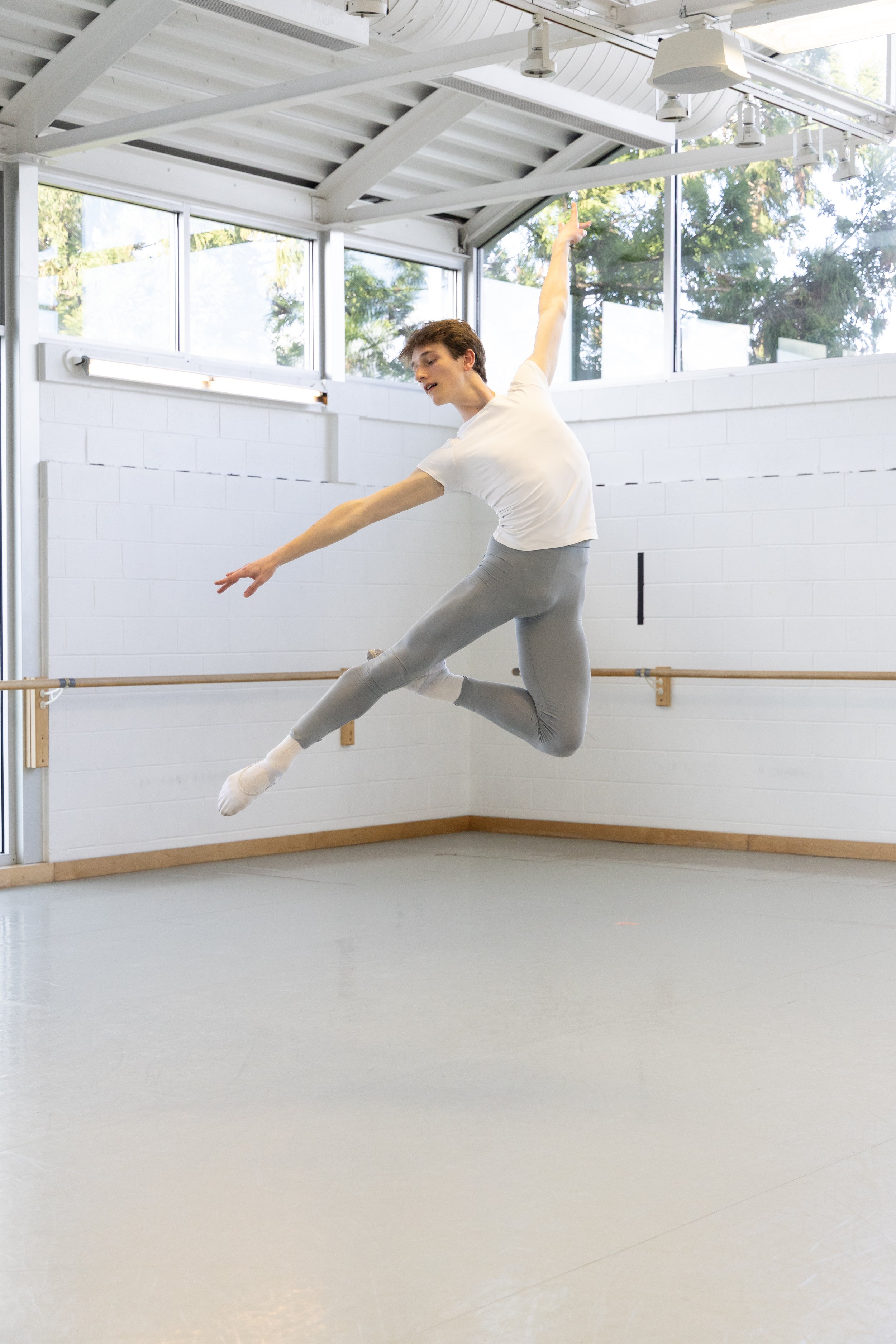 A male dancer captured mid-air during a dance move in a studio with white brick walls, large windows, and wooden barres.