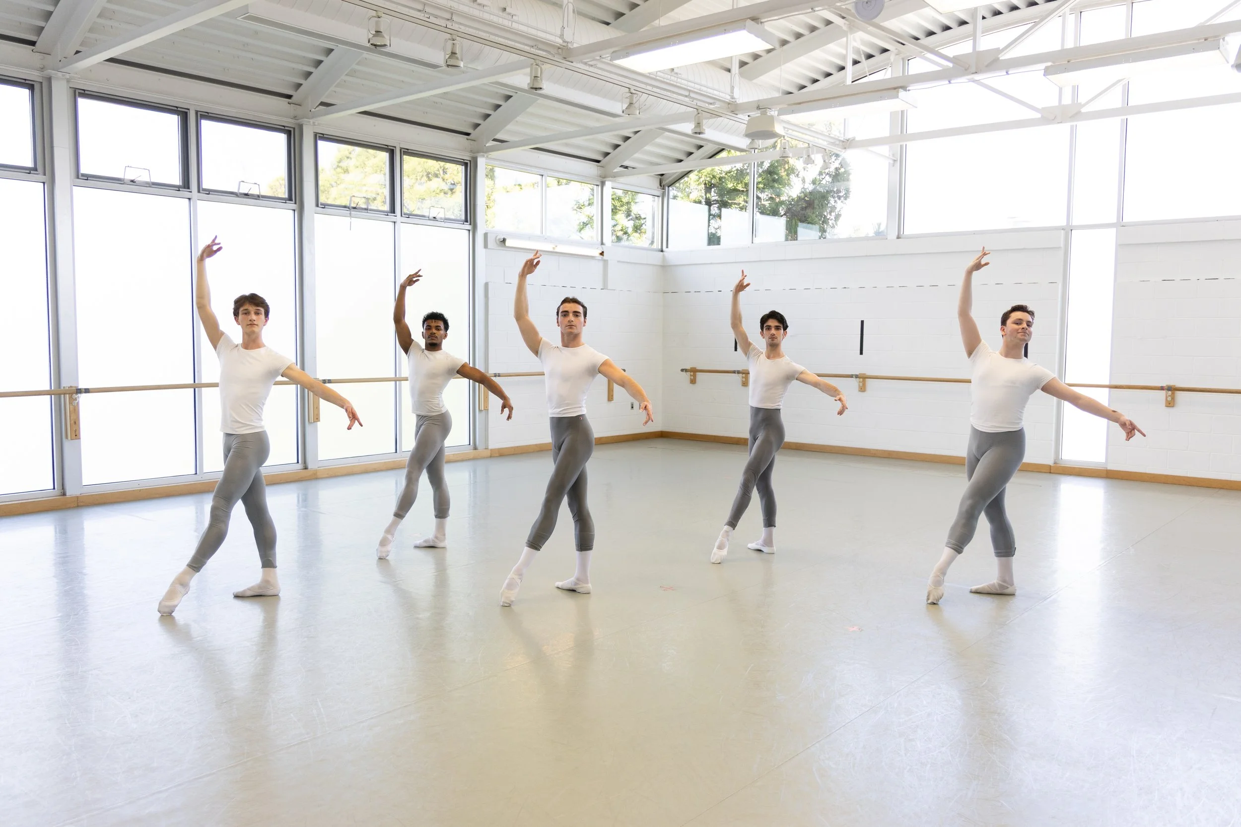 Five male ballet dancers practicing in a spacious dance studio with large windows and mirrored walls, wearing white t-shirts and gray leggings, performing ballet poses with arms raised and pointed toes.