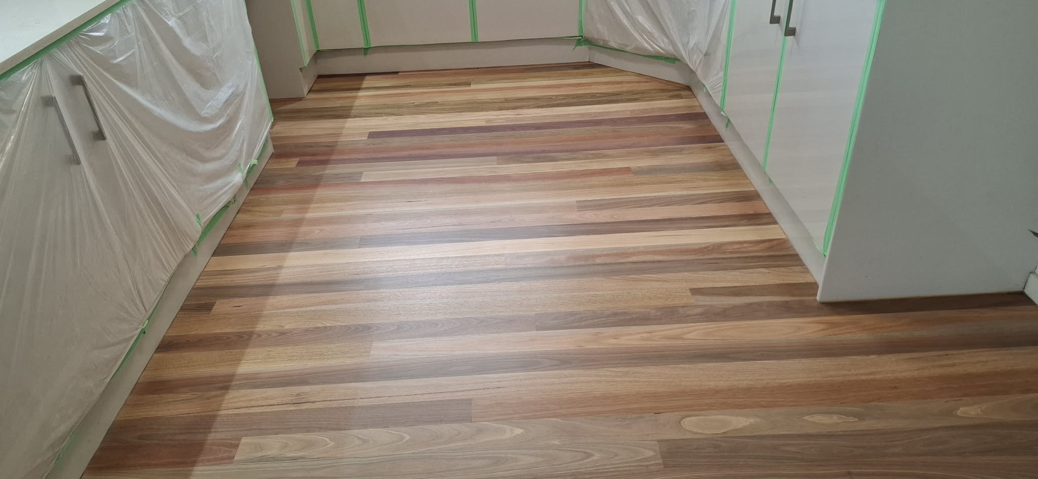 Wooden flooring in a kitchen with partially taped cabinets, indicating ongoing renovation or painting.