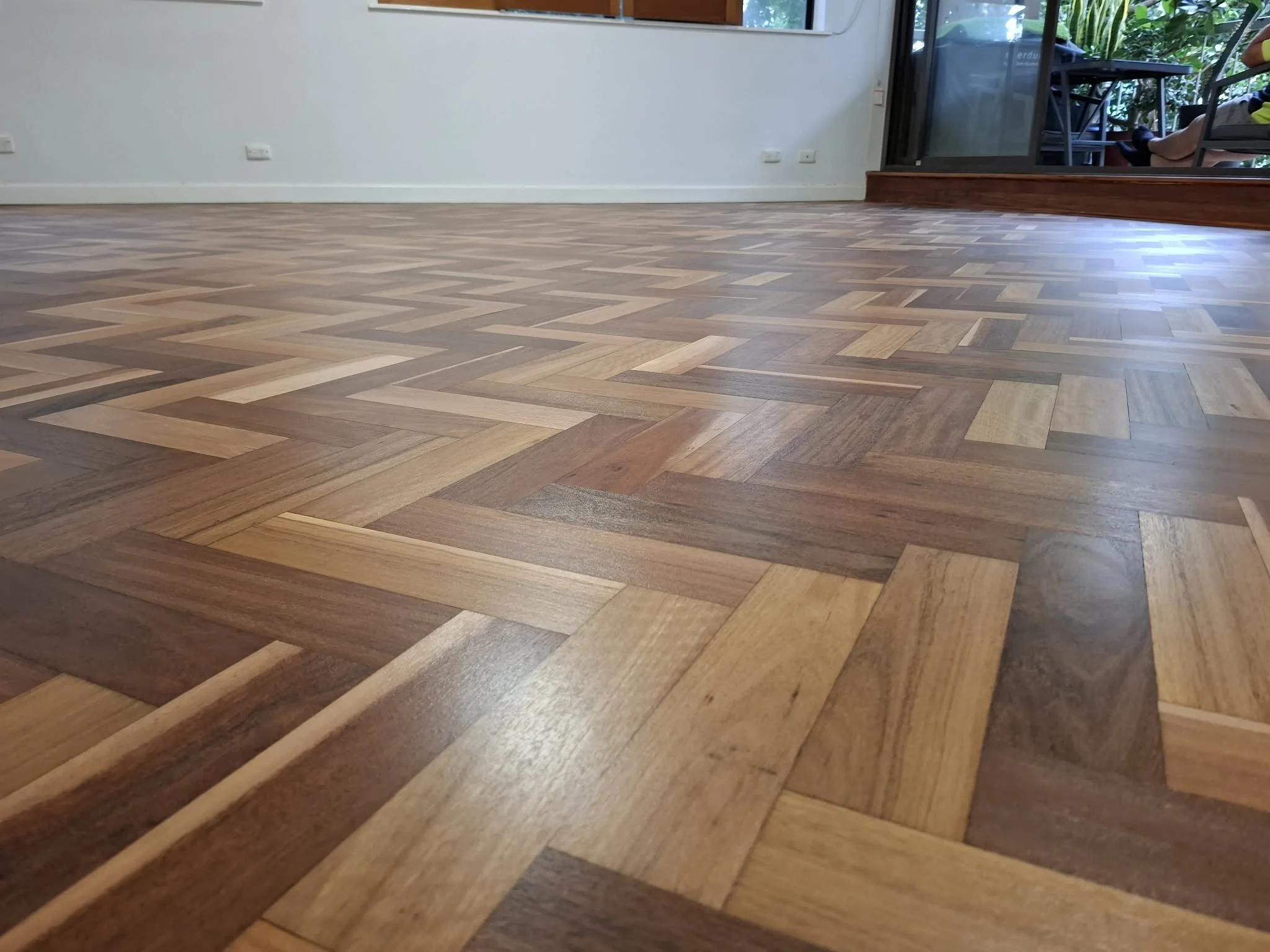 View of a room with a wooden parquet floor, white walls, and a sliding glass door leading to a balcony with outdoor furniture and greenery.