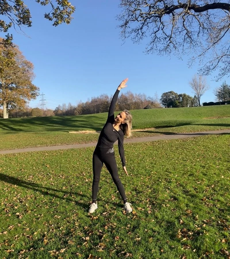 Hellen Barlow, Yoga for golfers coach, stretching outdoors on a grassy field with trees and clear blue sky in the background, showing a stretch movement for golfers.