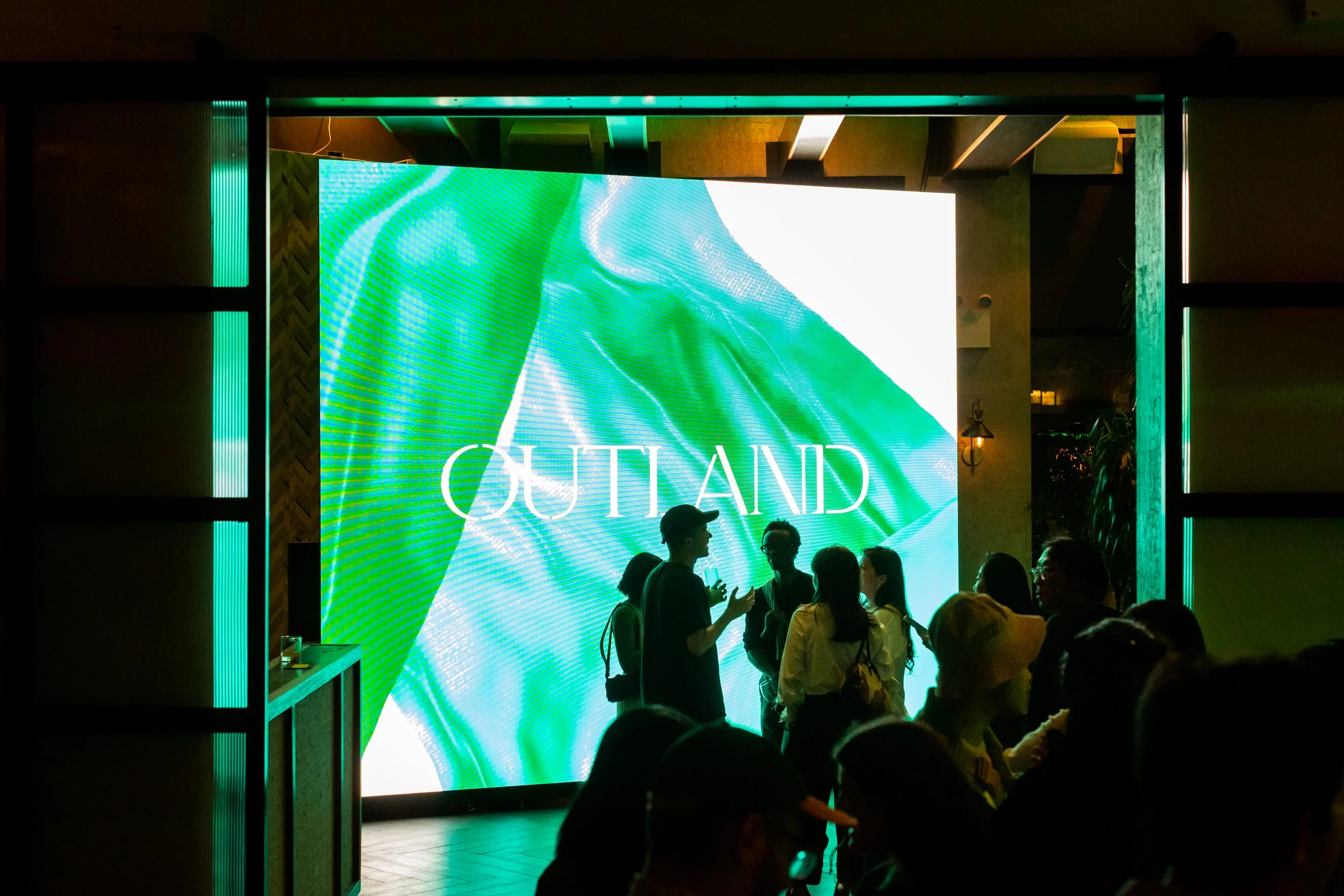 Group of people chatting in front of a large digital screen displaying the word 'OUTLAND' in a dimly lit, modern space.