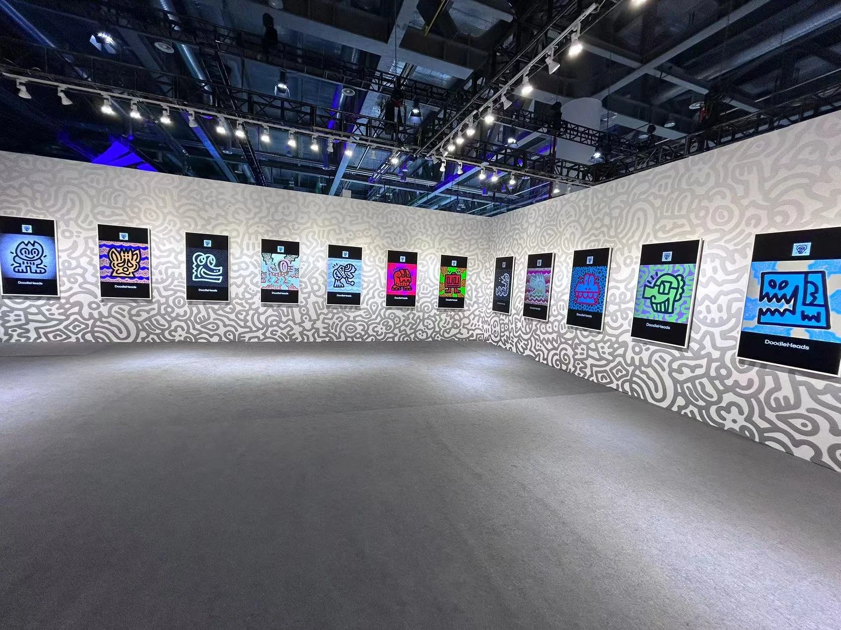 Art gallery exhibit featuring colorful digital artwork of doodle-style characters on wall-mounted screens in a modern, industrial space.