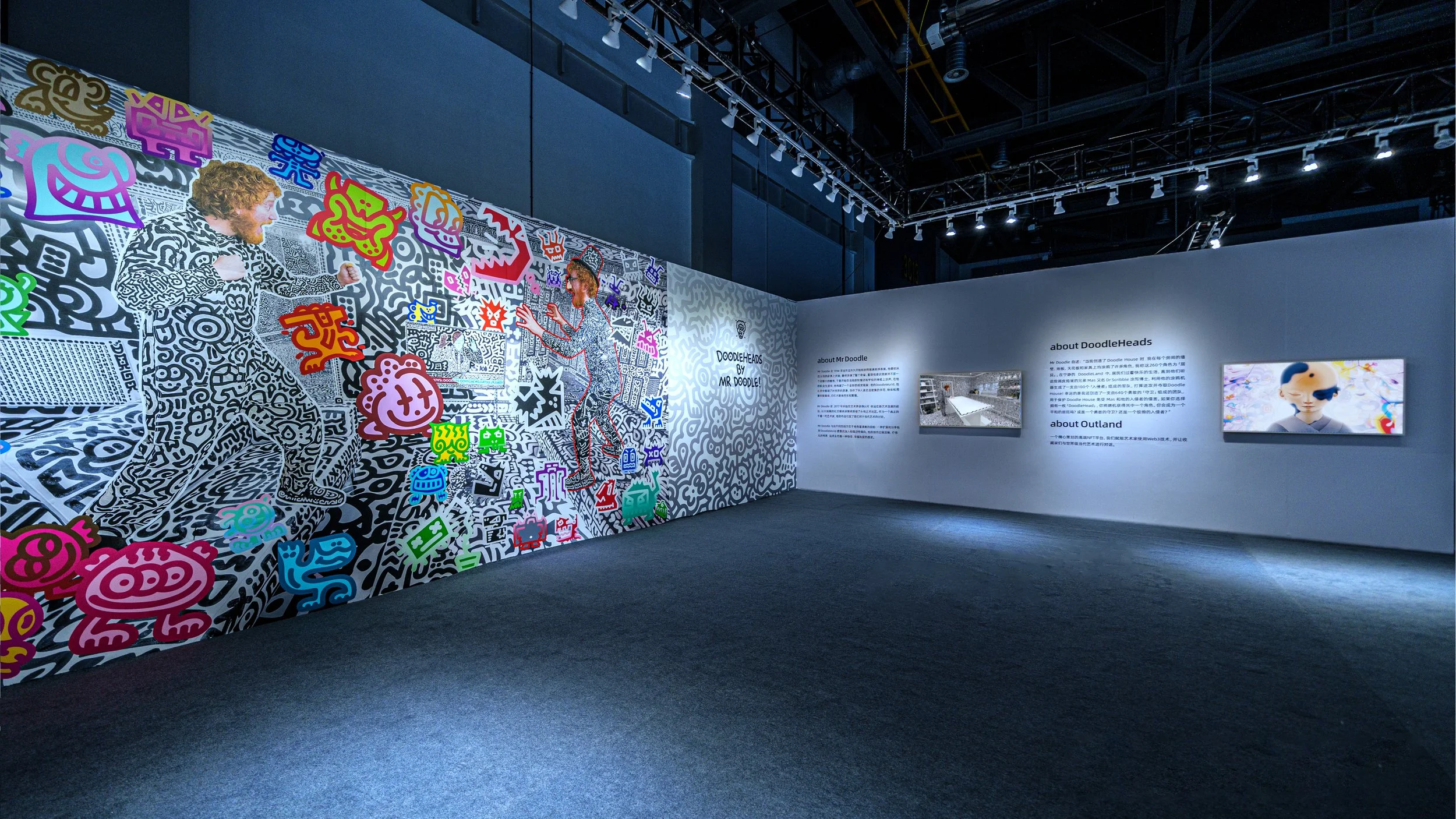Art exhibit featuring a mural with black and white patterns, colorful cartoon characters, and two people interacting. The setting appears to be an indoor gallery with informational panels on the wall.