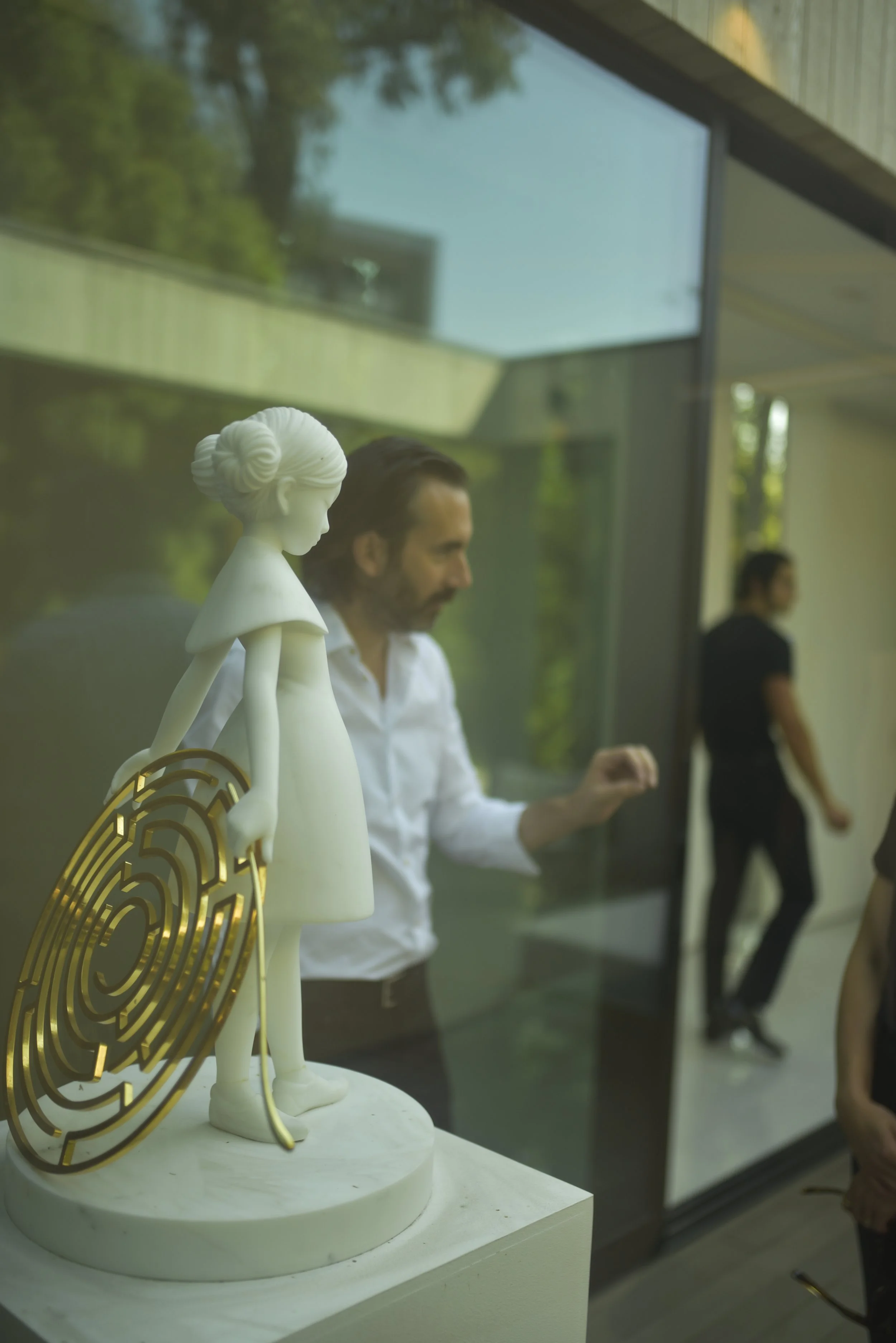 A white sculpture of a girl with a bun hairstyle holding a golden maze, placed on a pedestal, seen through a glass wall during an indoor event with two people in the background.