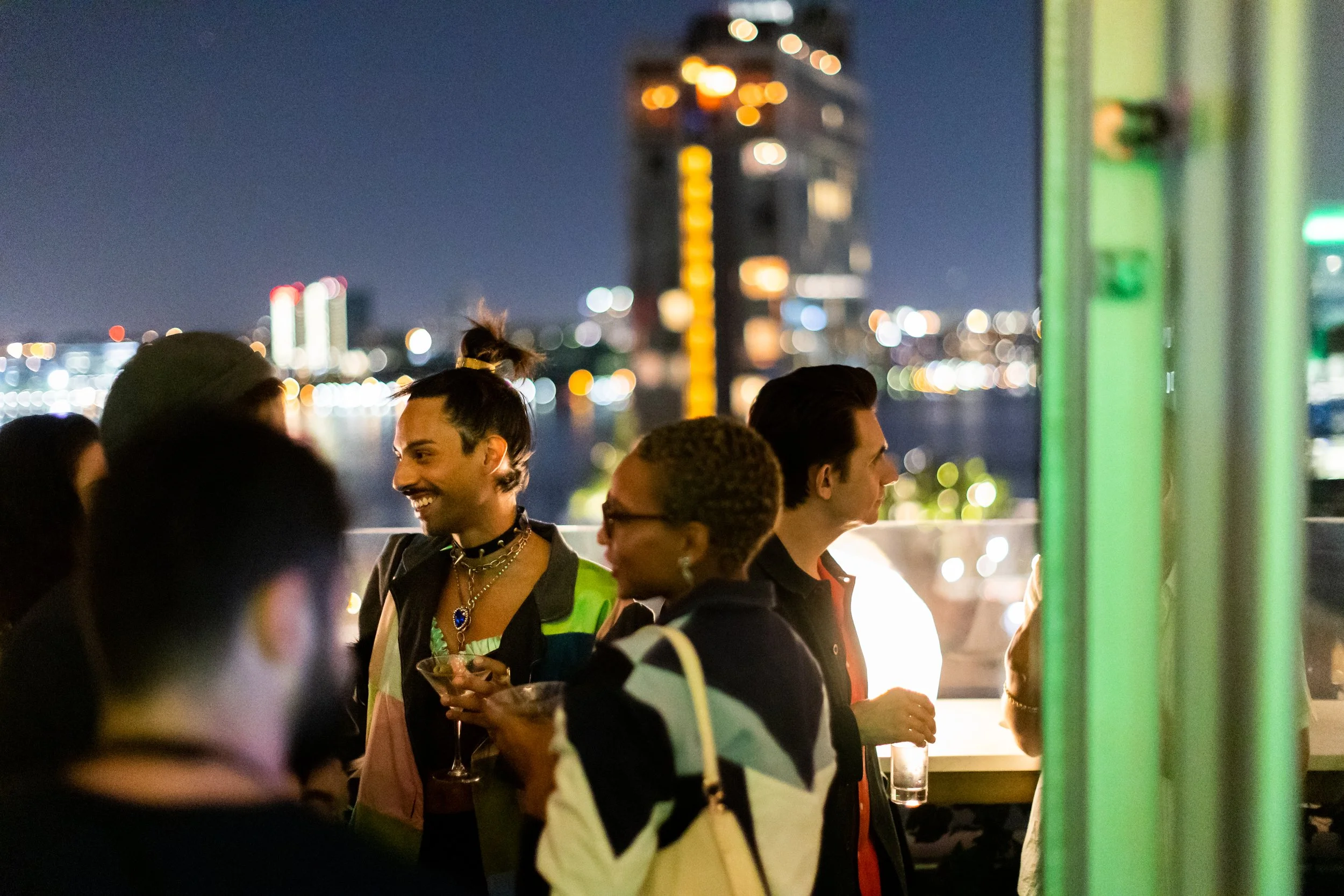 Group of diverse young adults socializing at night on a rooftop with a city skyline in the background. Some are holding drinks and talking, while others are looking out at the city lights.