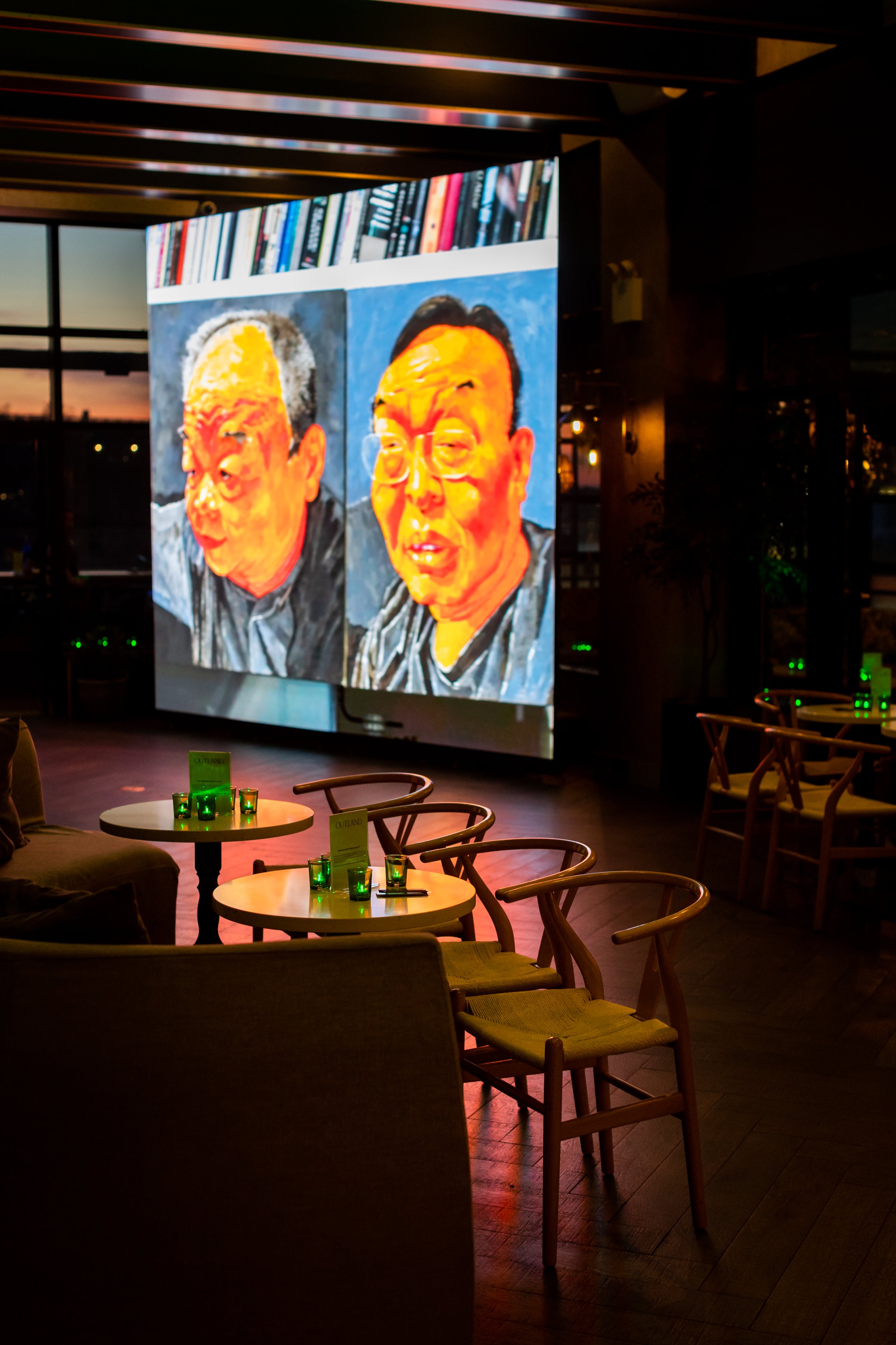 An indoor bar or lounge with a large digital screen displaying a painting of two men's faces, books on a shelf at the top of the screen, and green glass candle holders on tables.