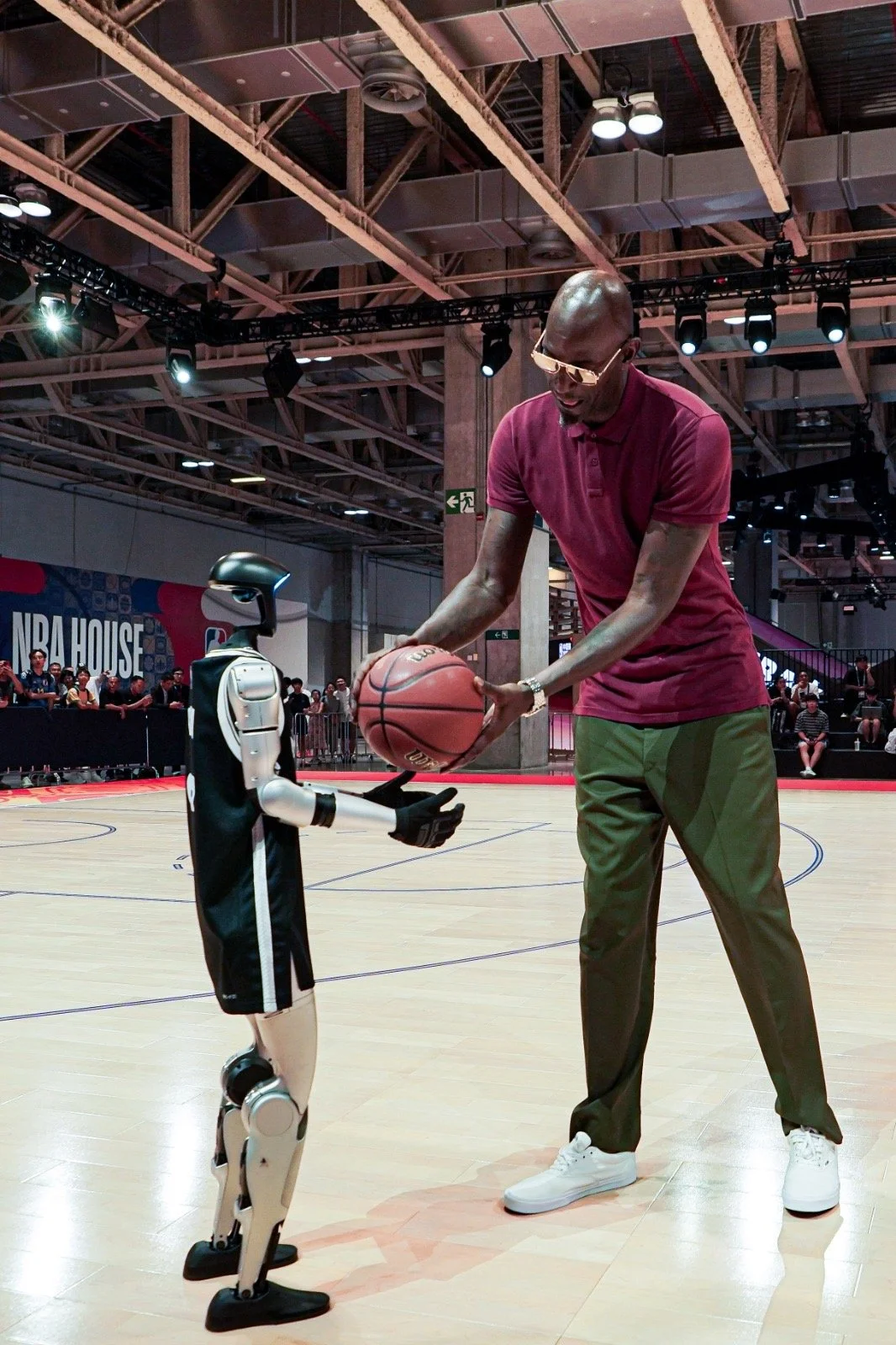 A tall man with glasses coaching a humanoid robot on playing basketball, passing a basketball on a court at NBA House.