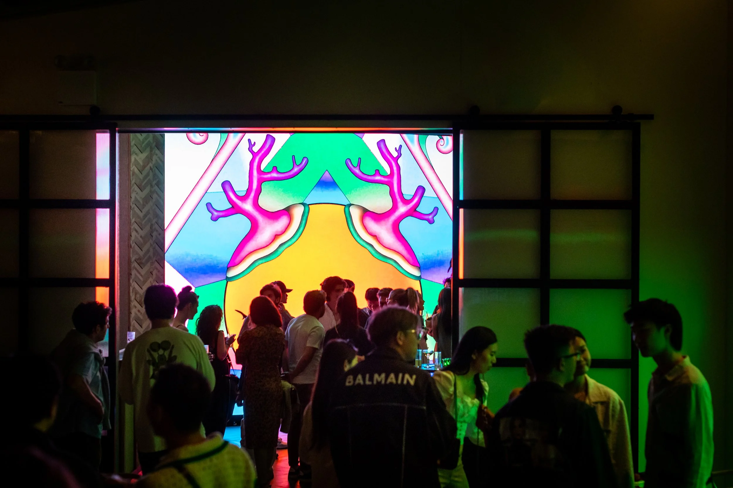 People gathering at an indoor event with a colorful, abstract digital artwork featuring pink branches and geometric shapes on a large screen in the background.