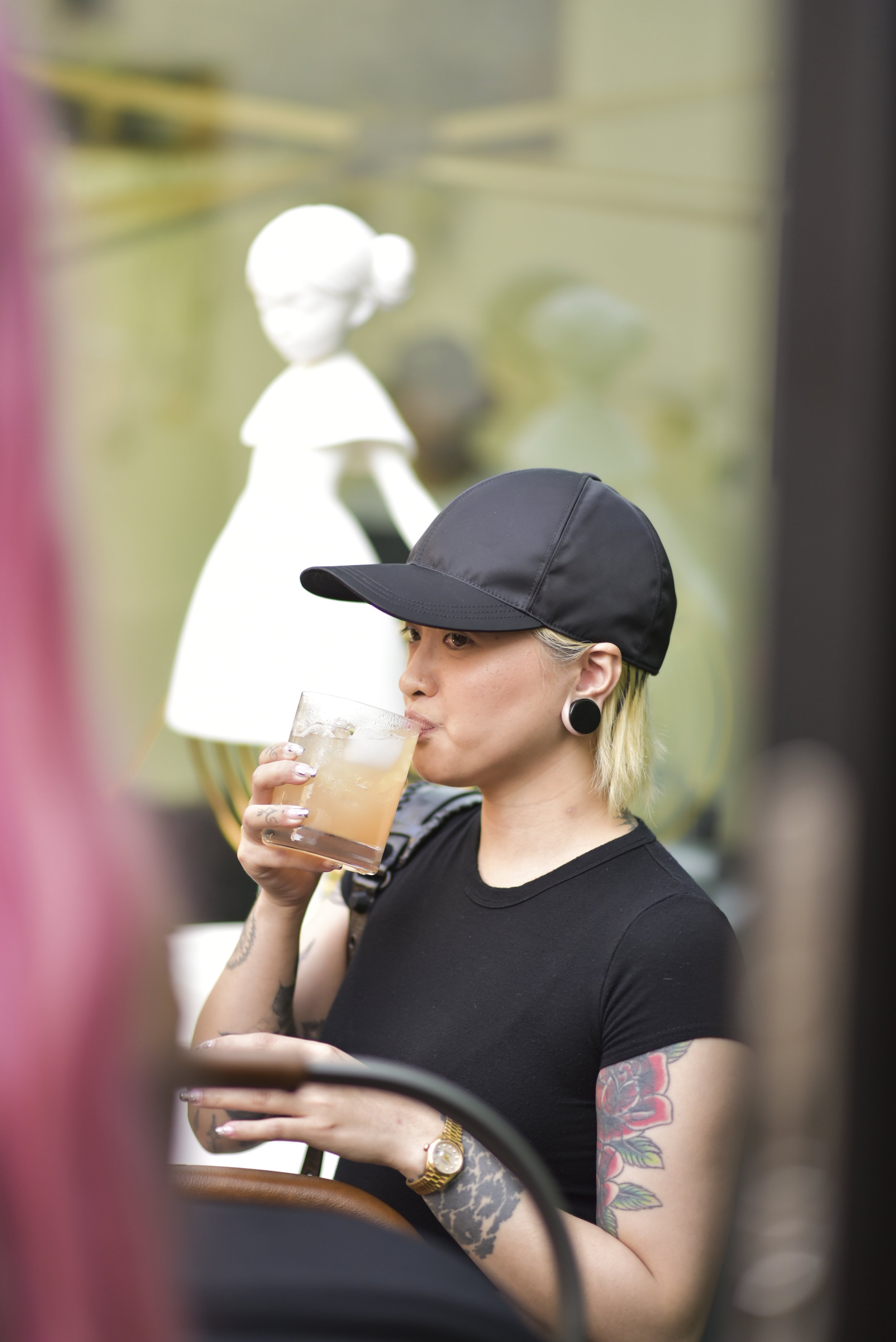 A woman with blonde hair, tattoos, and large earrings wearing a black cap and black t-shirt, drinking from a glass with ice while looking at her phone in a cafe or bar.