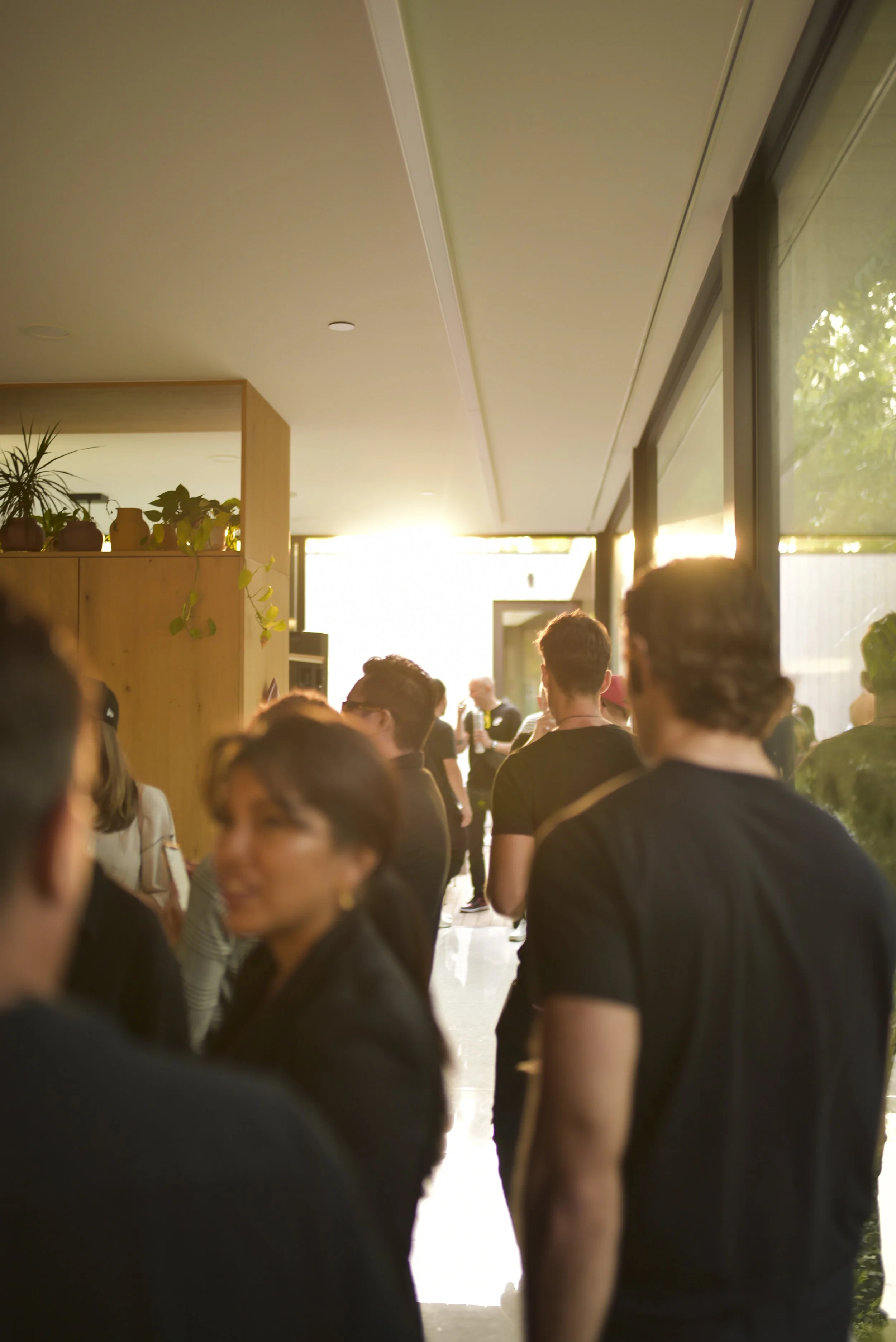 People standing in line indoors with sunlight shining in from the door, some having conversations.