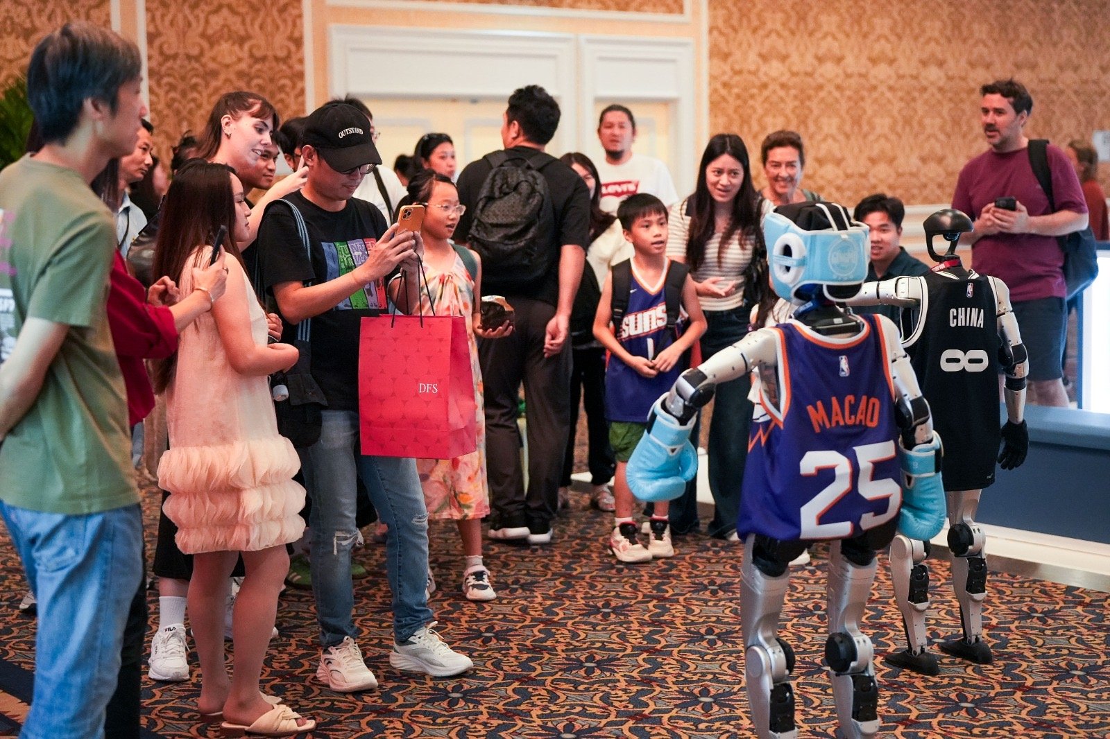 A group of people gathered around two robots dressed in basketball jerseys in a hotel lobby.