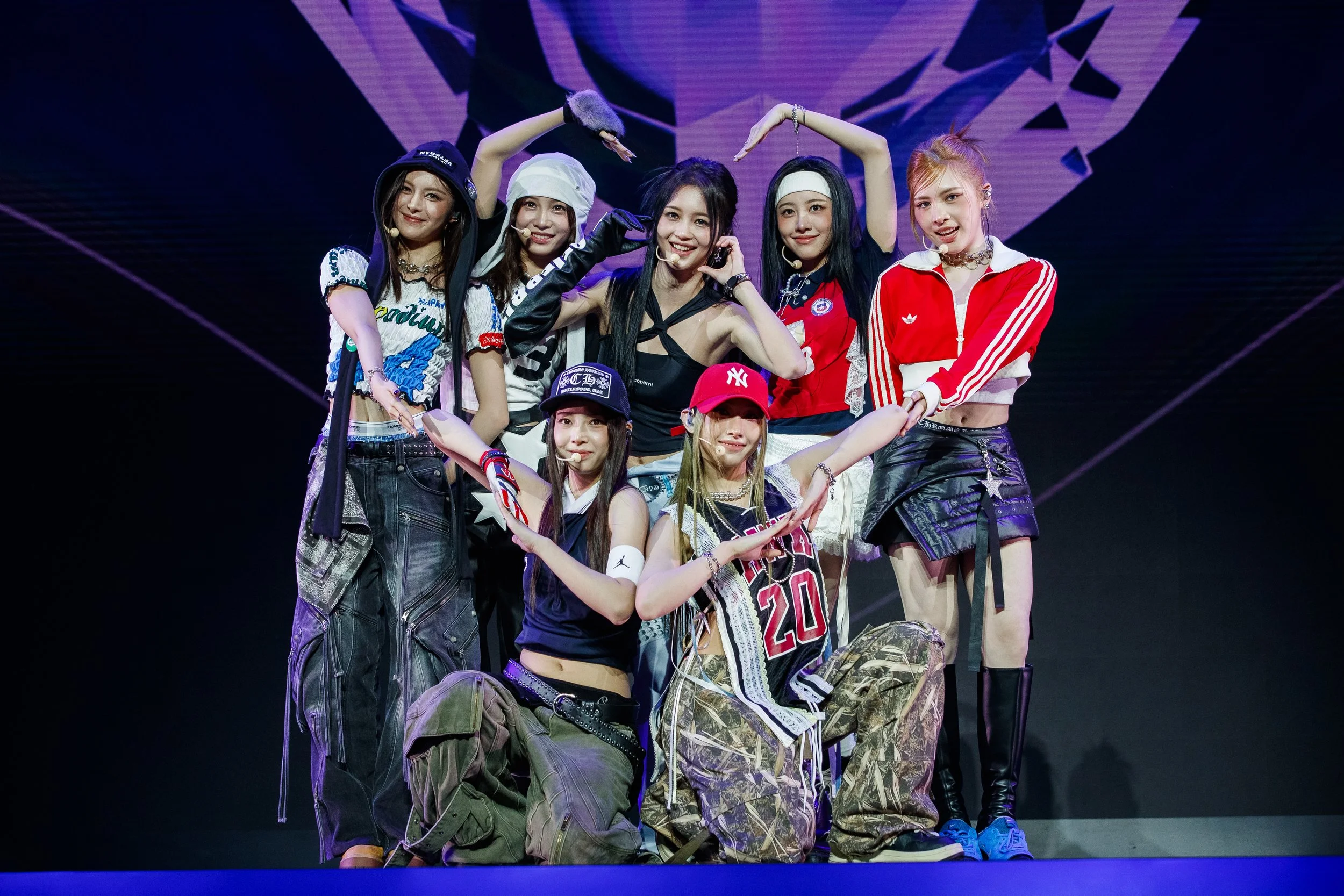 A group of eight young women in casual and sporty outfits, on a stage with a backdrop of colorful graphics. They are posing together, some making hearts with their hands and others smiling at the camera.