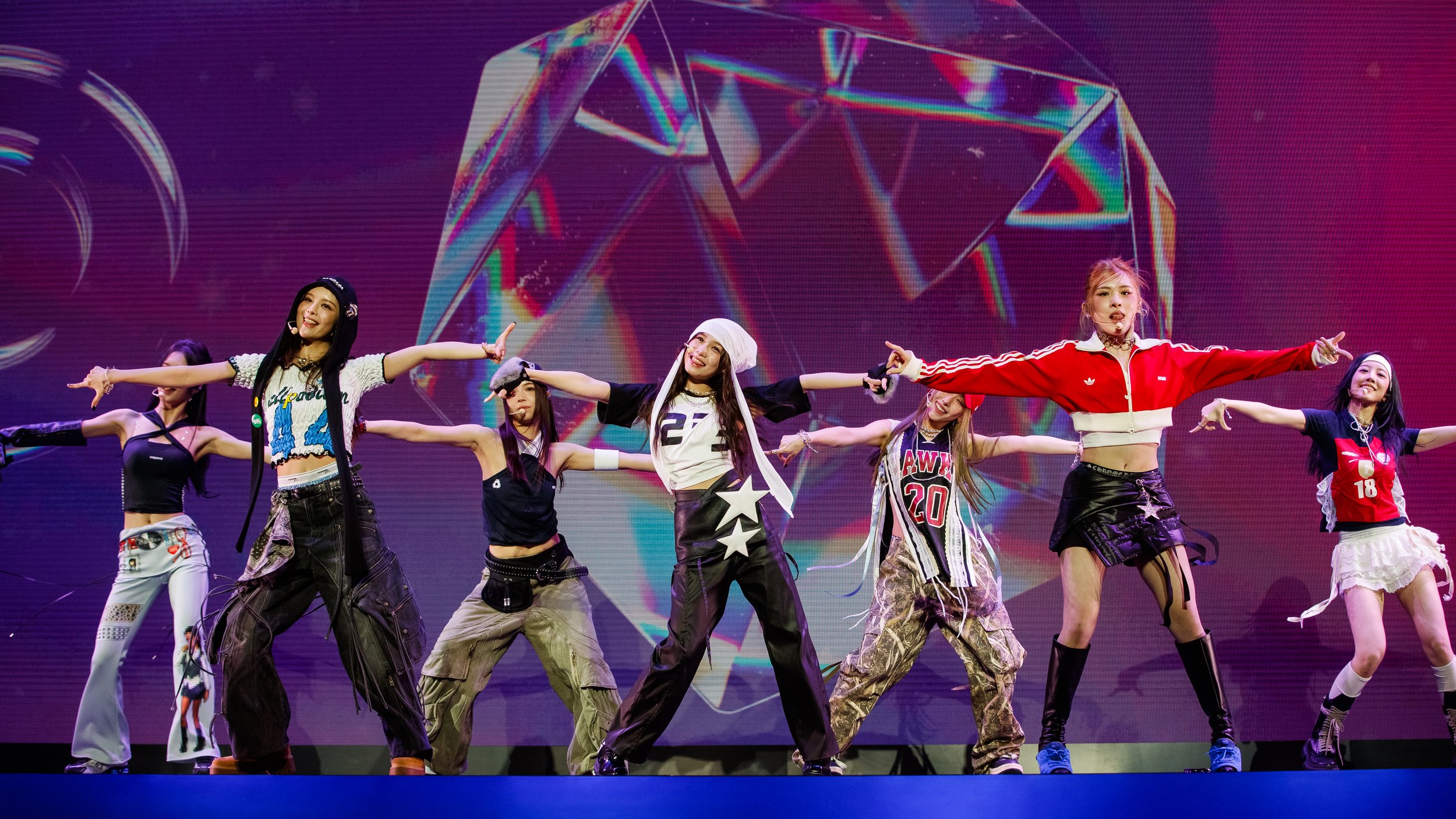 A group of seven young women are performing a dance routine on stage. They are dressed in colorful, casual, and sporty outfits, with some wearing crop tops, baggy pants, and accessories. A large digital screen with a crystal graphic design is in the 