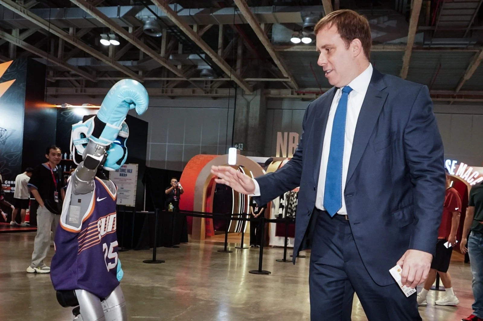 A man in a navy suit and blue tie high-fives a robot dressed in a Denver Nuggets basketball jersey and wearing a blue boxing glove at an indoor event.
