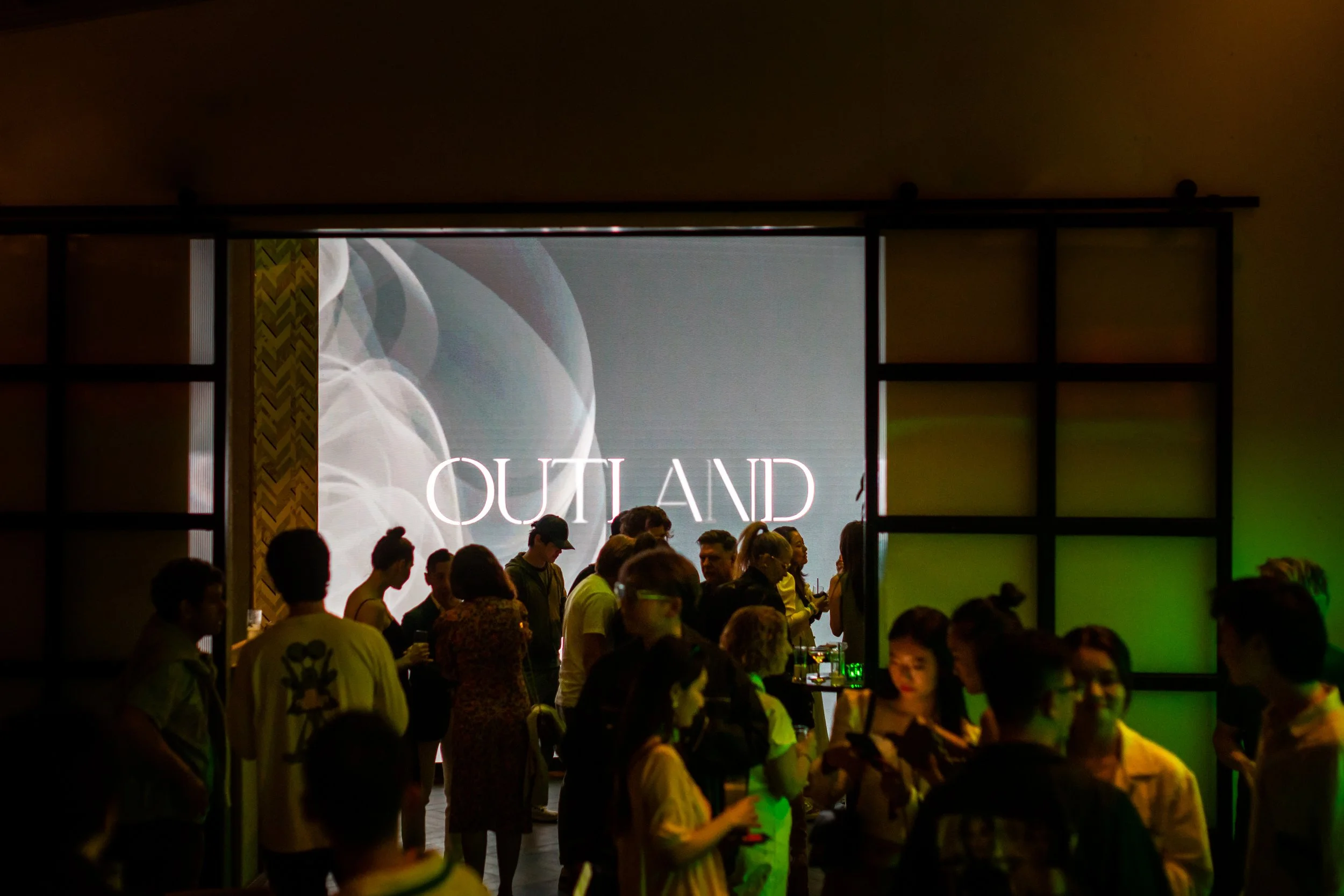 People gathered in front of a large screen displaying the word 'OUTLAND' at an indoor event.