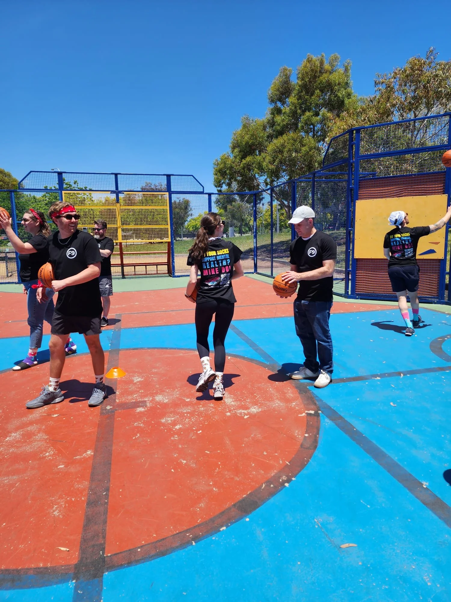 Group of people on a colorful outdoor basketball court, some holding basketballs, with trees and a clear blue sky in the background, participating in a Field9 Team Building experience in Adelaide.