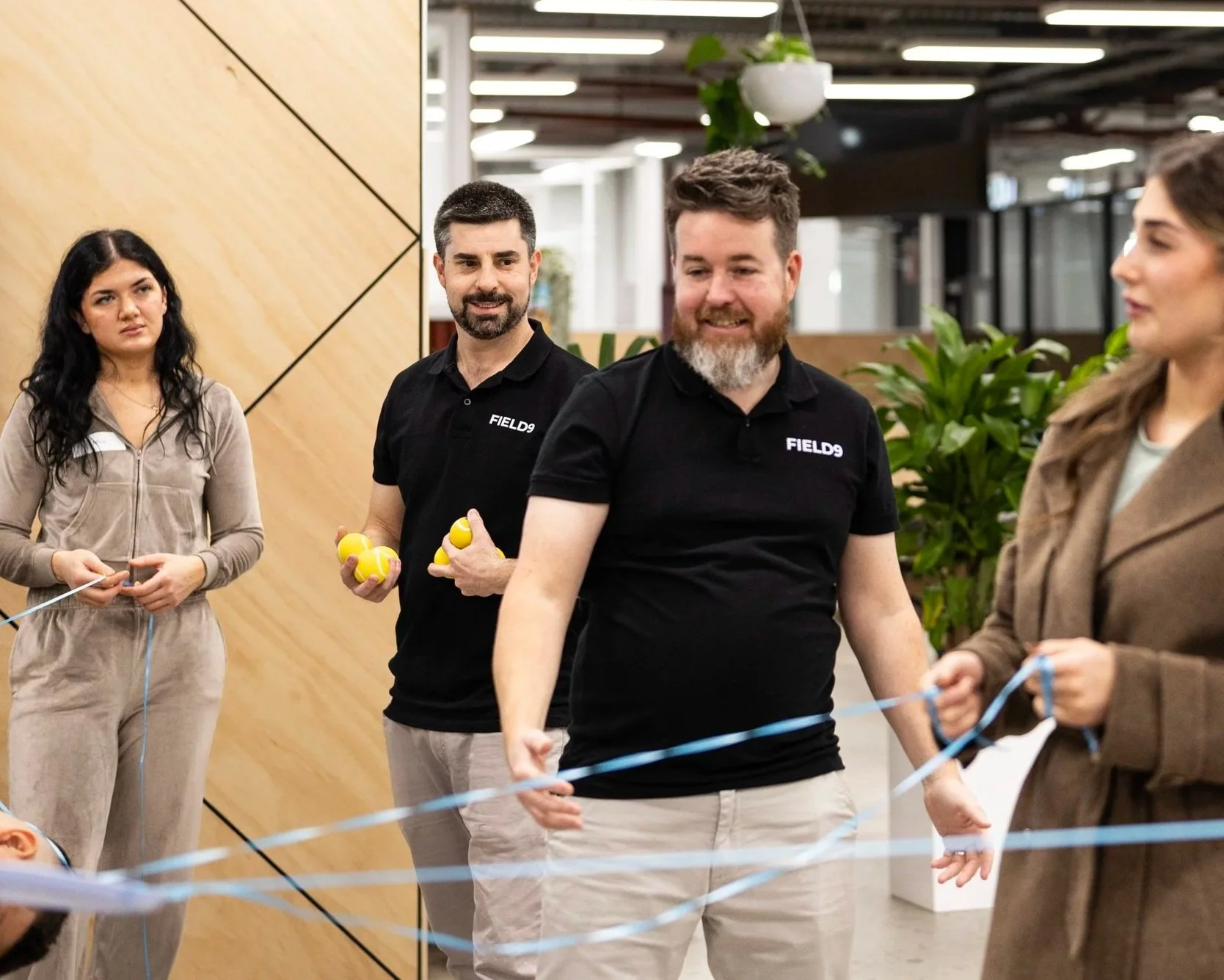 Group of people holding yellow balls, participating in a team-building activity hosted by Field9 in Adelaide, in a modern office environment with plants and wooden wall decor.