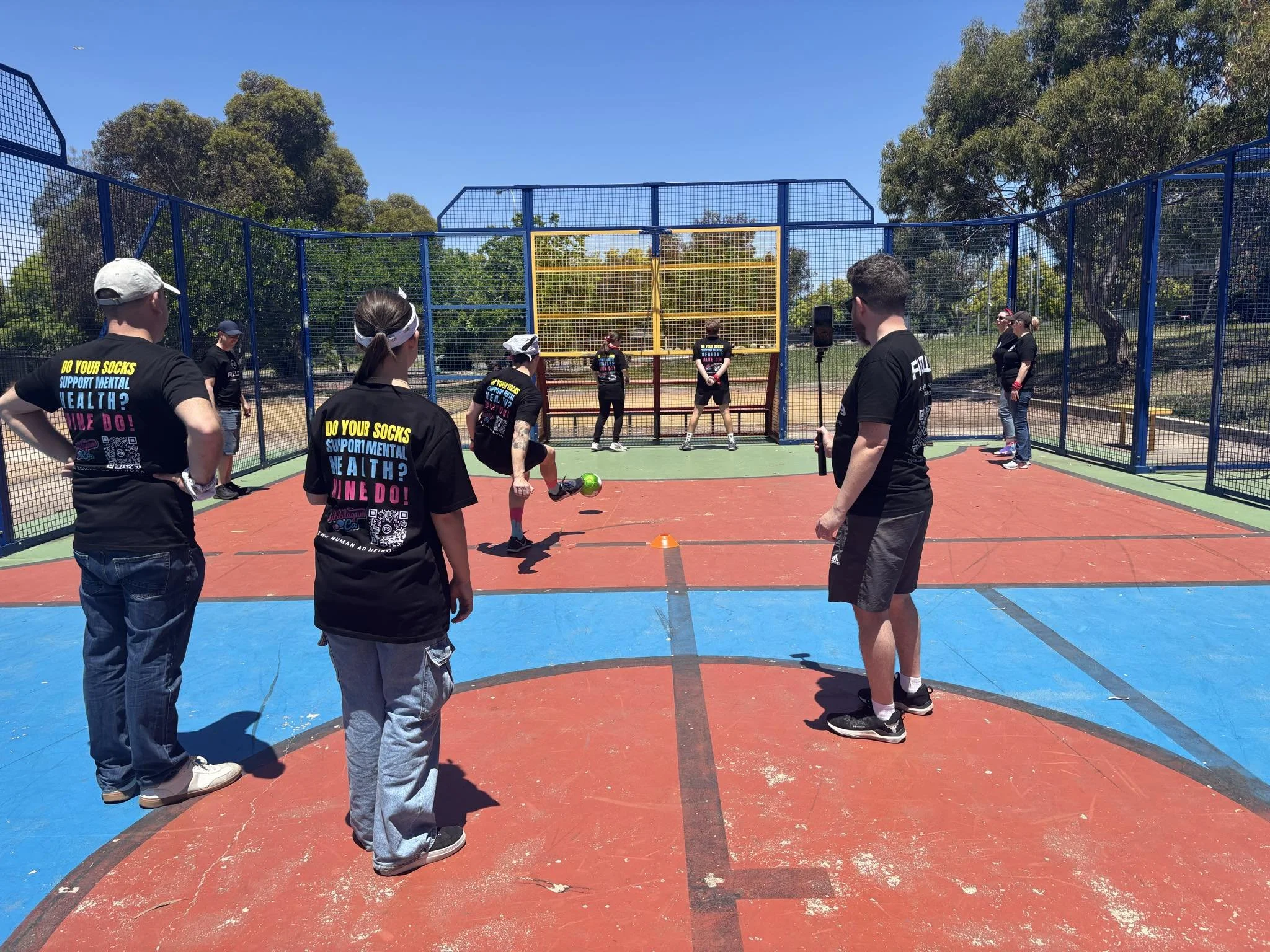 A group of people playing soccer inside a fenced court on a sunny day, with trees in the background, participating in a Field9 Team Building experience in Adelaide.
