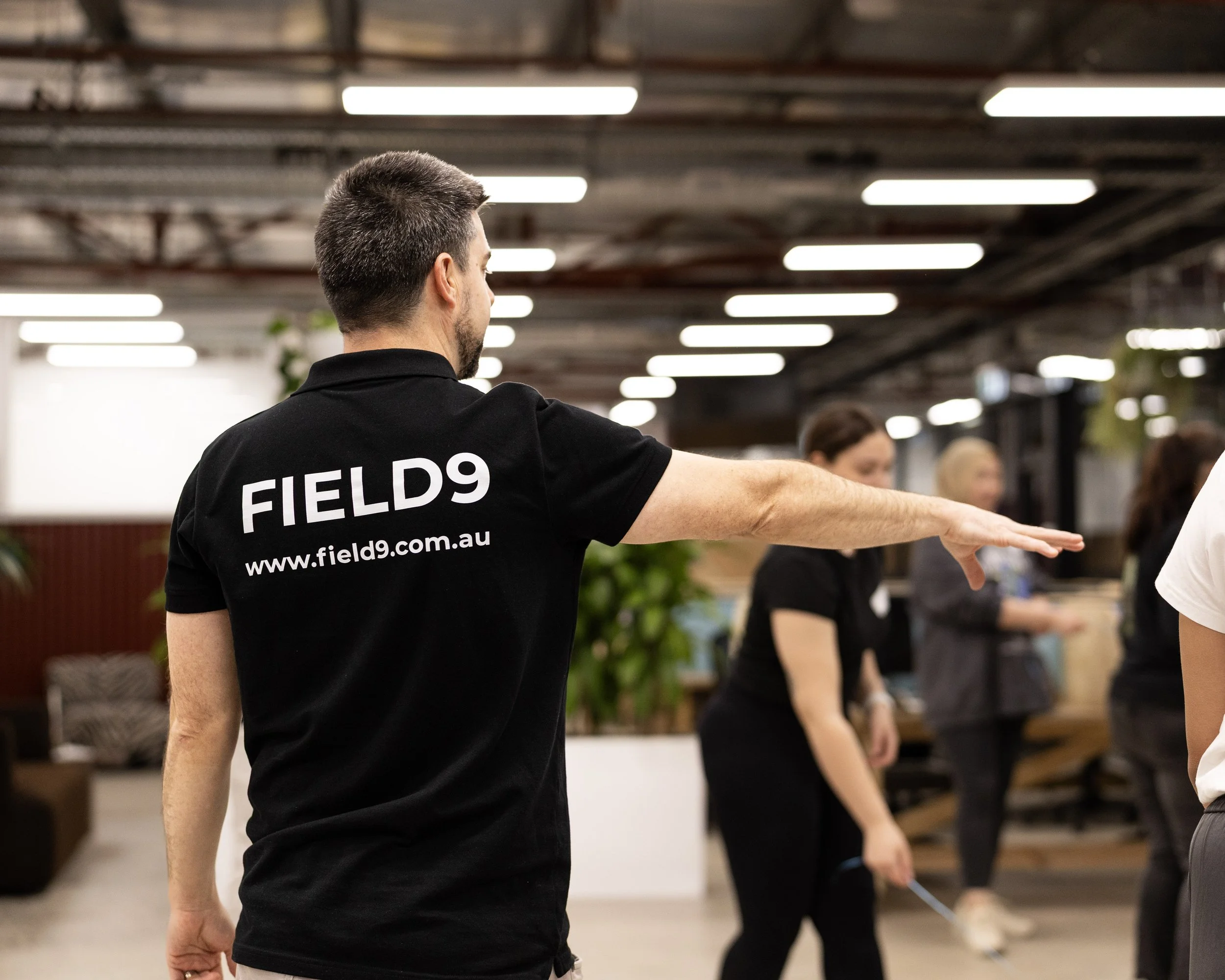 A man with short dark hair wearing a black shirt with the words 'FIELDS9' and a website URL on the back, standing in an indoor setting with fluorescent lighting, leading a team building activity in Adelaide.