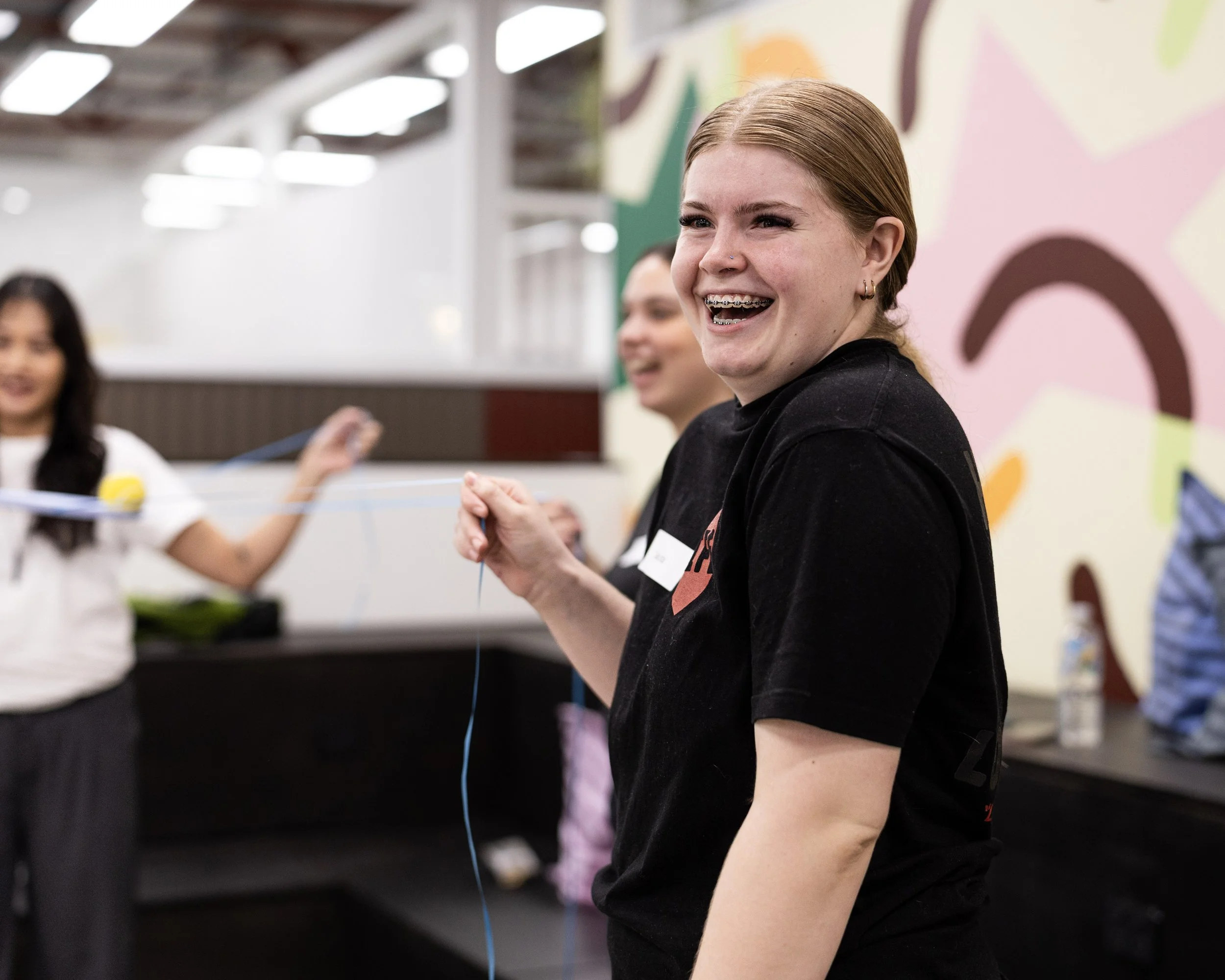 A young woman with tattoos on her arm, wearing braces and a nose piercing, smiling while holding a blue string or thread at an indoor event, with other people and colorful wall art in the background, enjoying a Field9 team building experience.