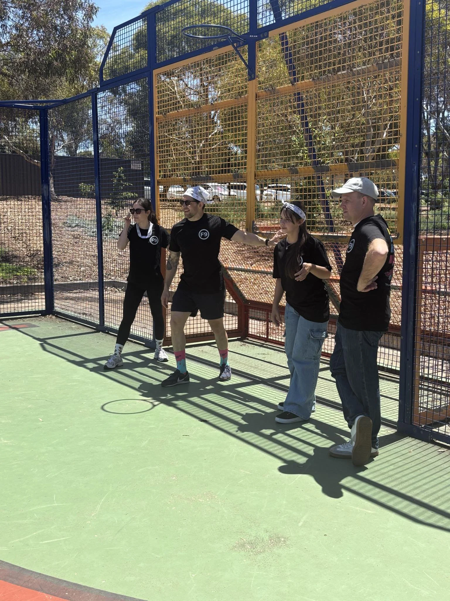 Four people standing on an outdoor court next to a high metal fence, with trees and parked cars in the background, engaging in a game of pickleball.