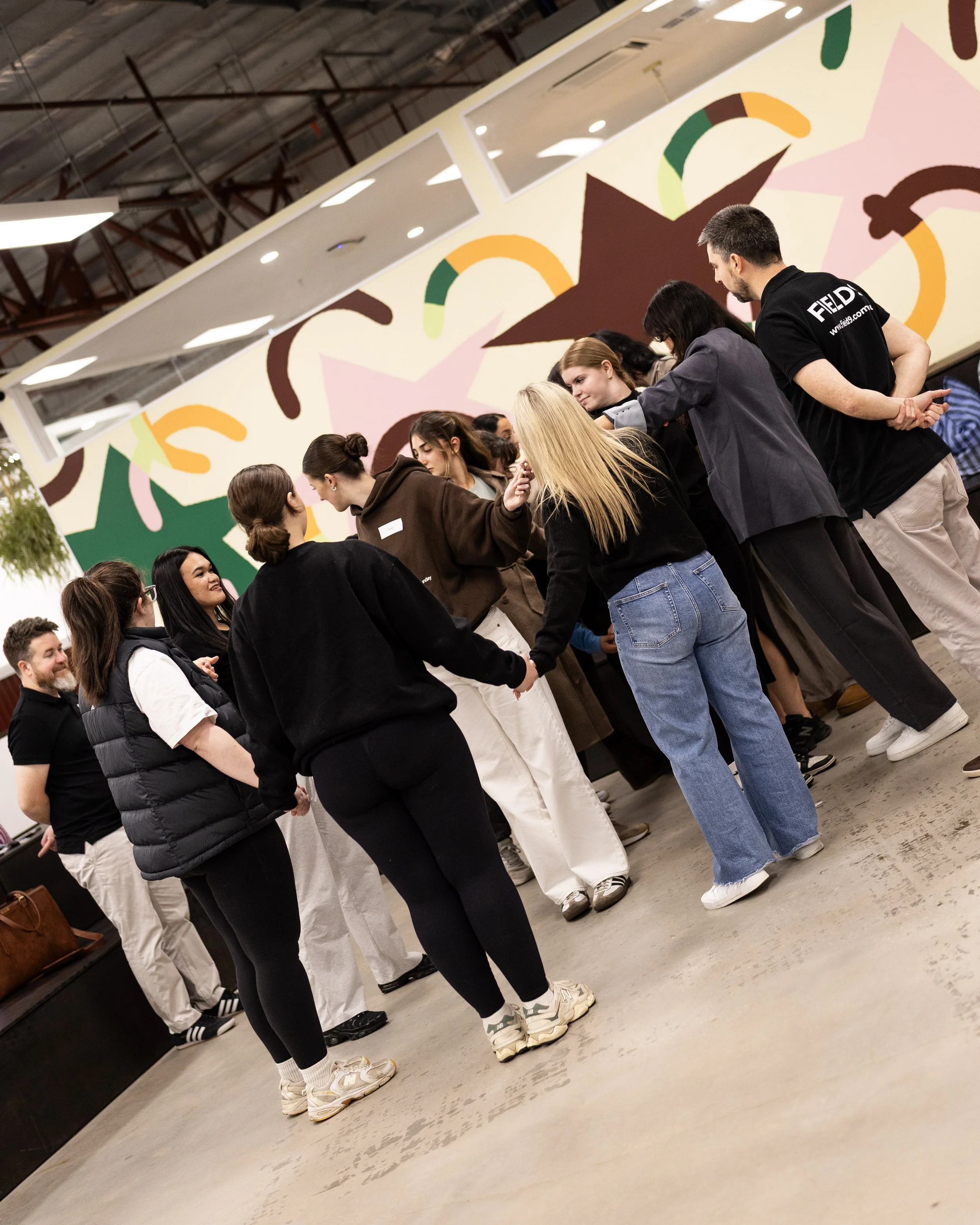 Group of people standing in line, holding hands, inside a building with colorful wall art featuring stars and rainbows, engaging in a Field9 team building experience in Adelaide.