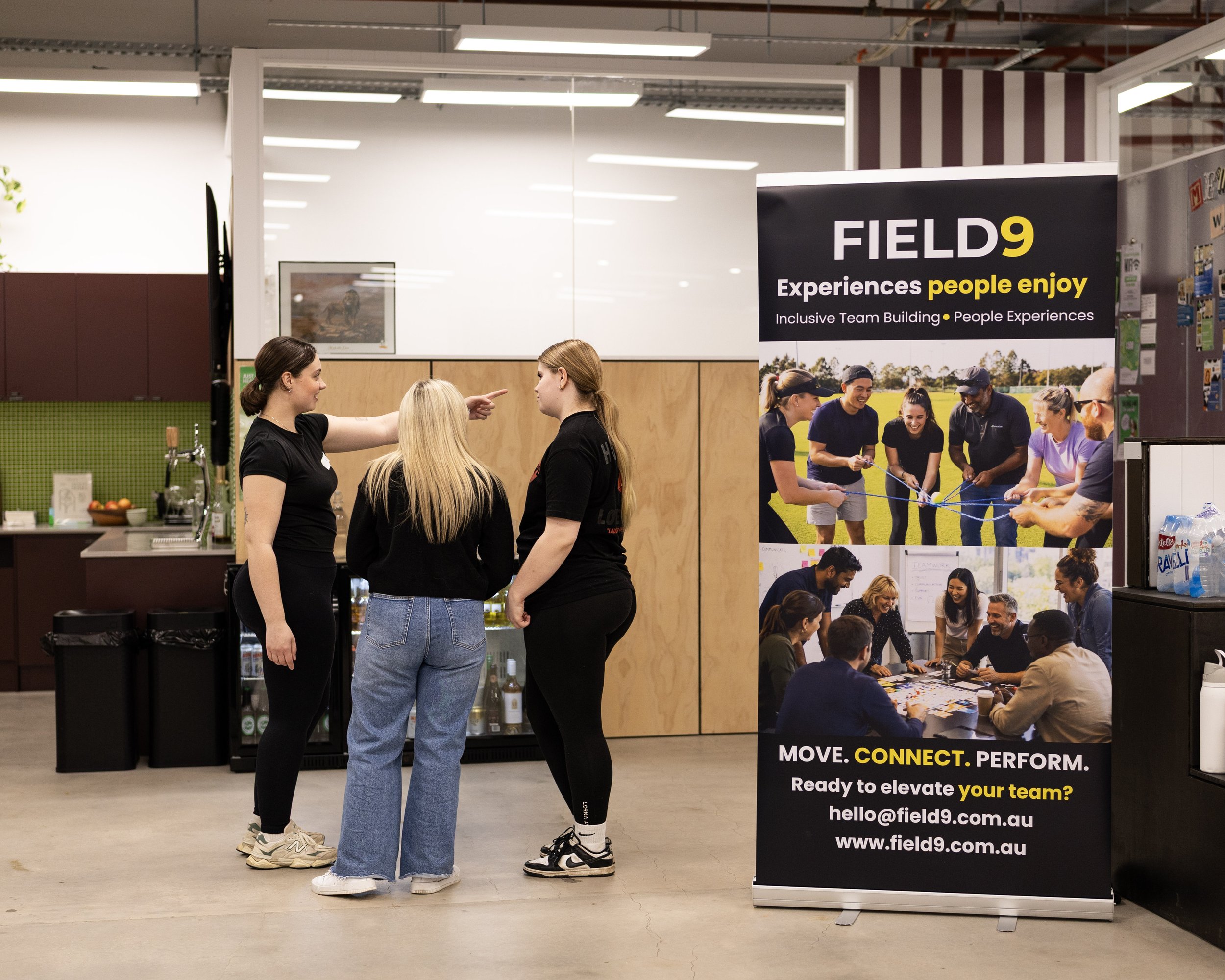 A team attending a Field9 session, standing near a display board with colourful images and text promoting team building activities. The display board reads 'FIELD9 Experiences people enjoy. MOVE. CONNECT. PERFORM. Ready to elevate your team?'