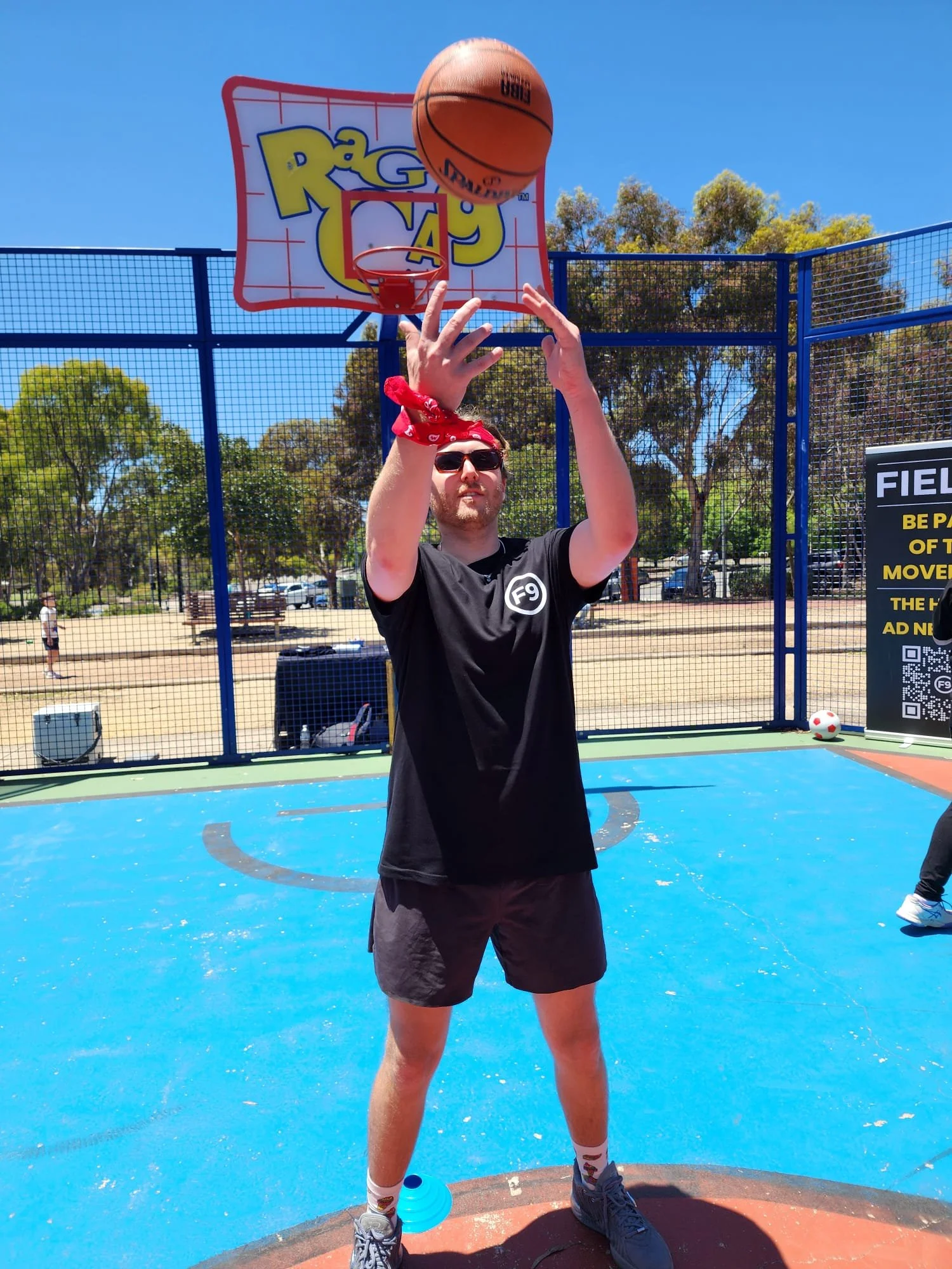 A man wearing sunglasses, a red bandana, a black T-shirt, black shorts, and sneakers stands at a baseball field, holding a basketball above his head near a basketball hoop, with trees and a parking lot in the background on a clear, sunny day.