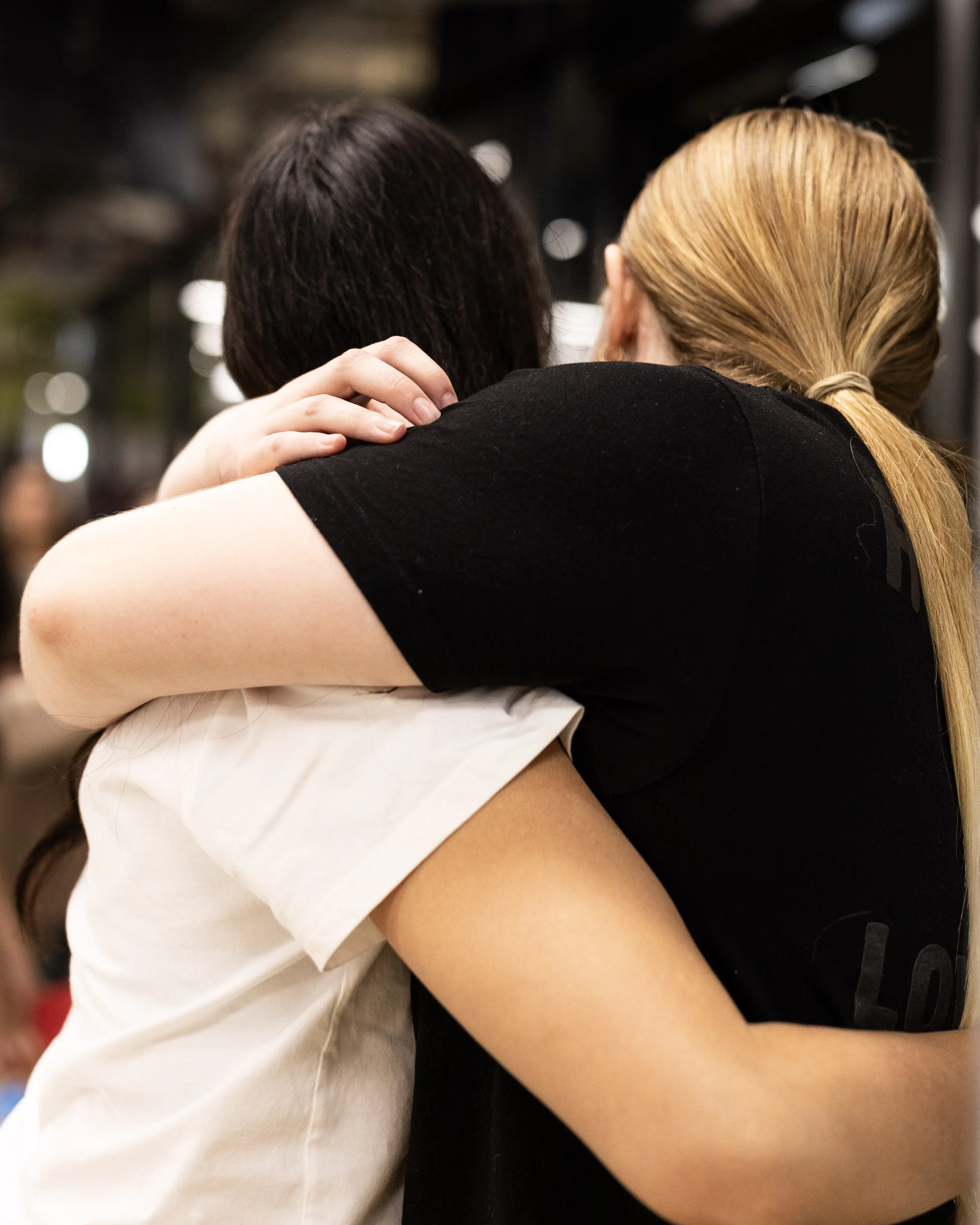 Two people hugging, one with dark hair and one with blonde hair in a ponytail, at a Field9 team building experience in Adelaide, with blurred lights in the background.
