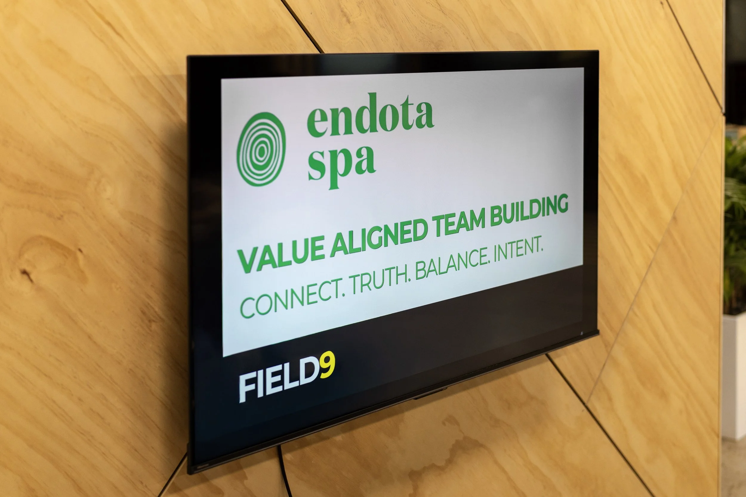 A digital screen showing the logo and name of Endota Spa, with the text 'Value Aligned Team Building' and words 'Connect. Truth. Balance. Intent.' in green, hosted by Field9 in Adelaide. The background is a wooden wall.