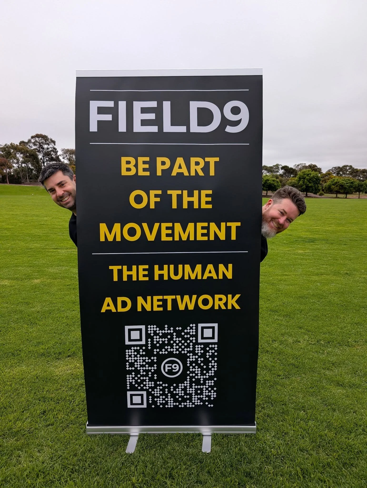 Two men peeking from behind a black event sign on a grassy field, the sign reads "FIELD9 BE PART OF THE MOVEMENT THE HUMAN AD NETWORK" with a QR code at the bottom.