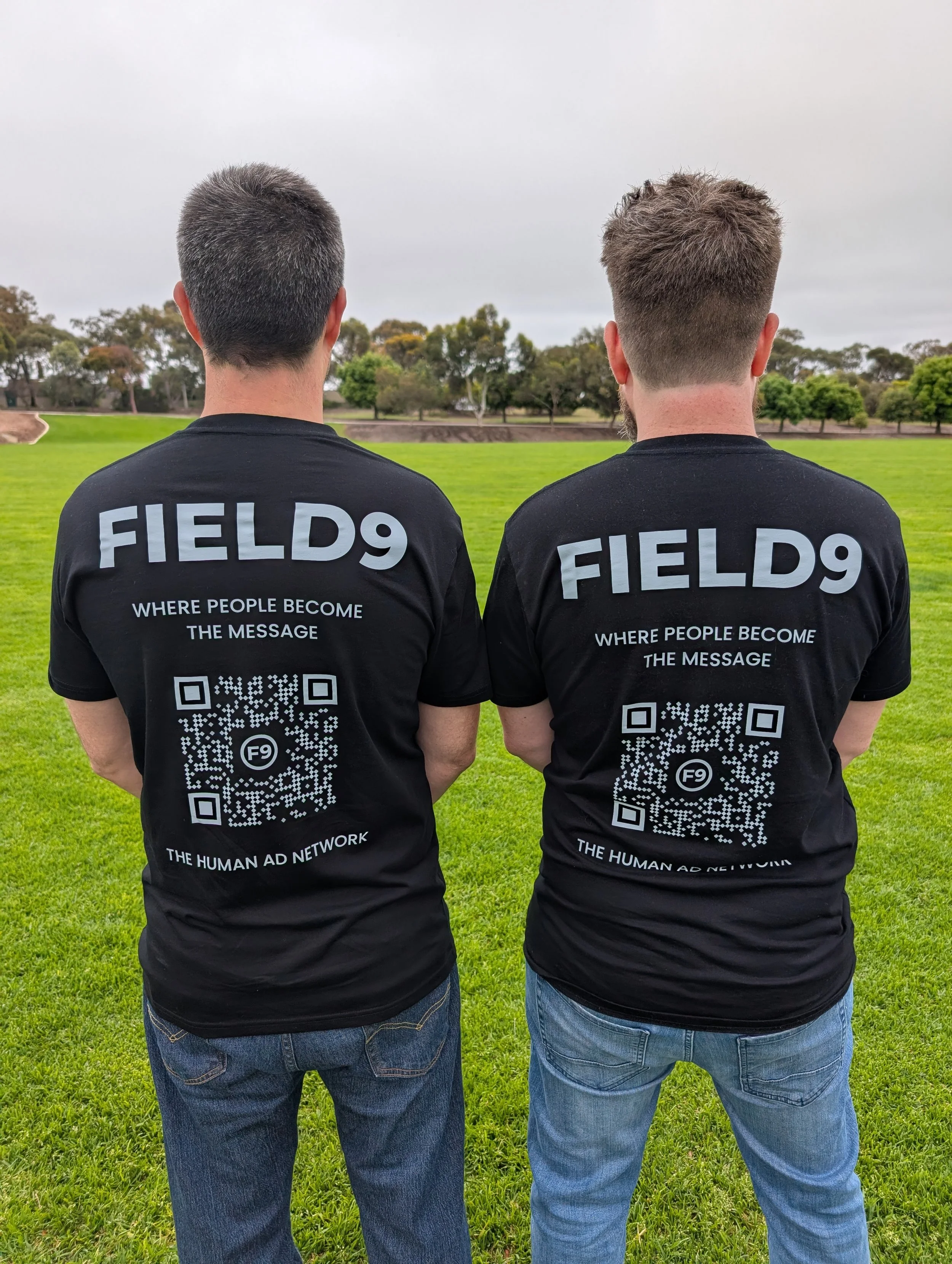 Two men standing on a grassy field wearing black t-shirts with 'FIELD9' printed on the back, featuring QR codes and the text 'WHERE PEOPLE BECOME THE MESSAGE,' 'F9,' and 'THE HUMAN AD NETWORK.'