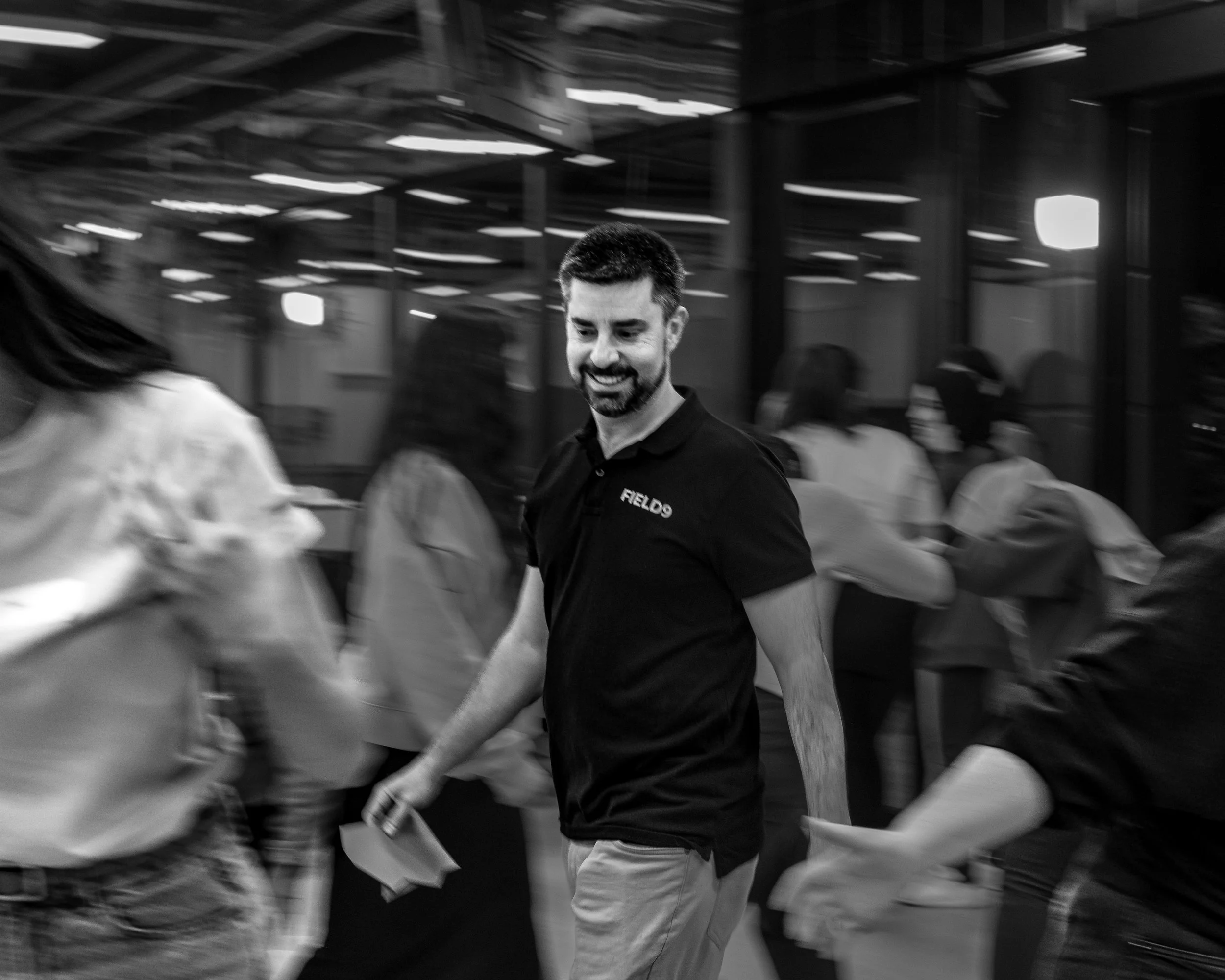 A Field9 facilitator smiling and walking in a crowded indoor team building event in Adelaide, with mirror walls and ceiling lights.
