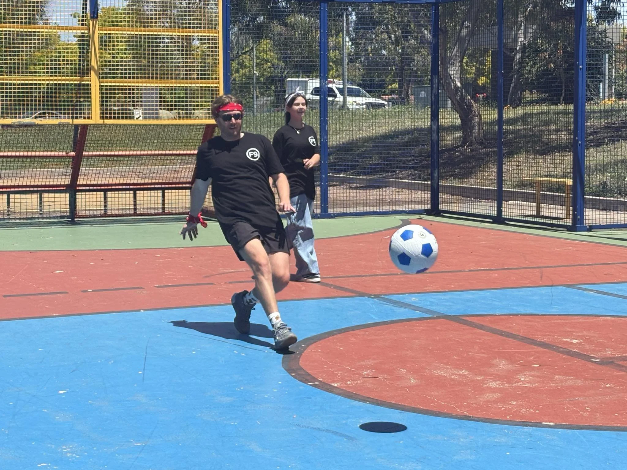 A young man in black athletic clothing, sunglasses, and a red bandana is playing soccer on an outdoor court with red and blue markings. A young woman in similar attire is watching or waiting nearby. The court is surrounded by a colorful metal fence w