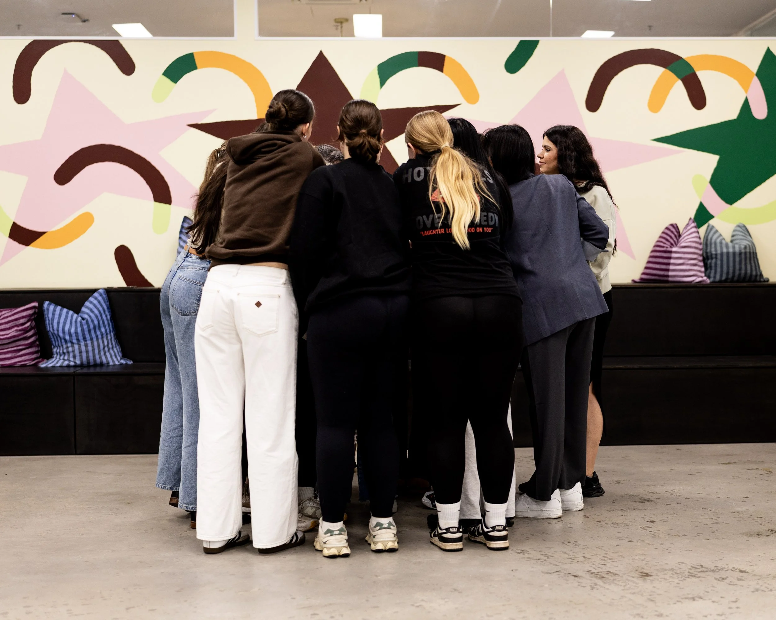 A group of eight young women are standing in a circle indoors, facing each other. They are dressed casually in jeans, sweatpants, and jackets, with some wearing sneakers. They are gathered in front of a colorful abstract mural on the wall, which feat
