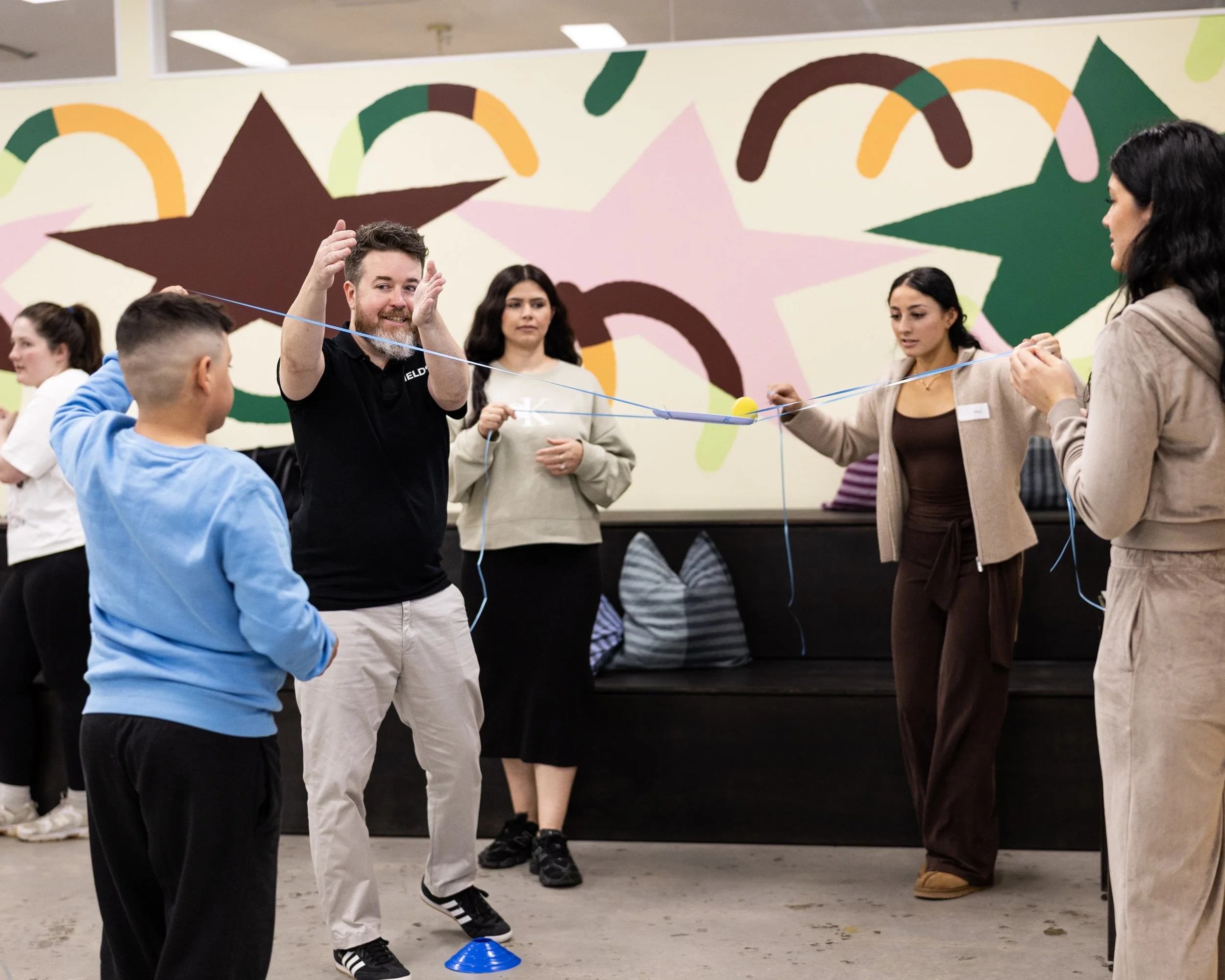 Group of diverse people participating in Field9nteam-building activity, standing in a circle, holding a string with a yellow ball in the center, indoors with colorful wall art in the background.
