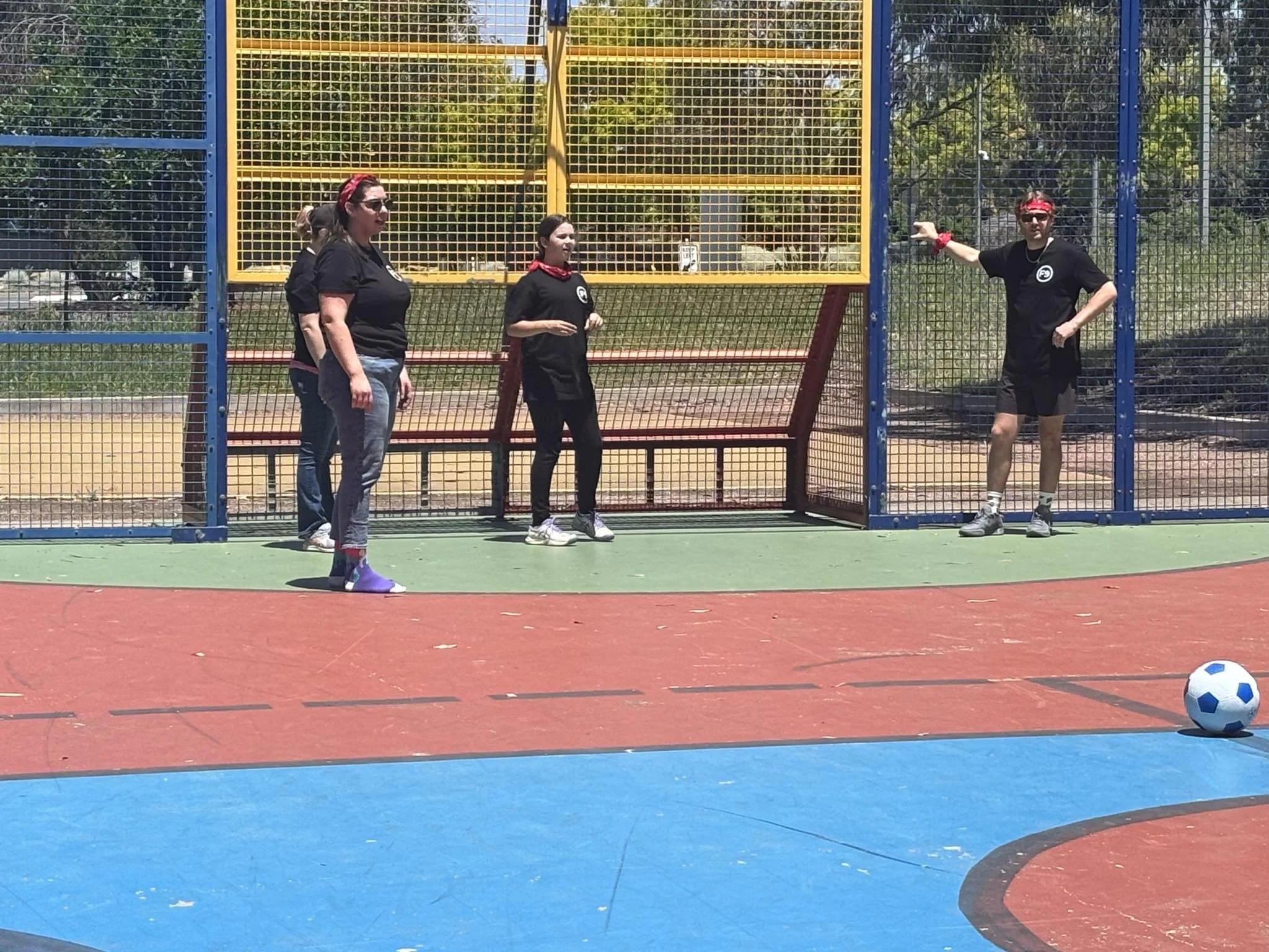 Group of four women standing on a tennis court, two of whom are wearing black shirts and red bandanas, with a soccer ball in the foreground and a colorful metal fence behind them, participating in a Field9 Team Building experience in Adelaide.