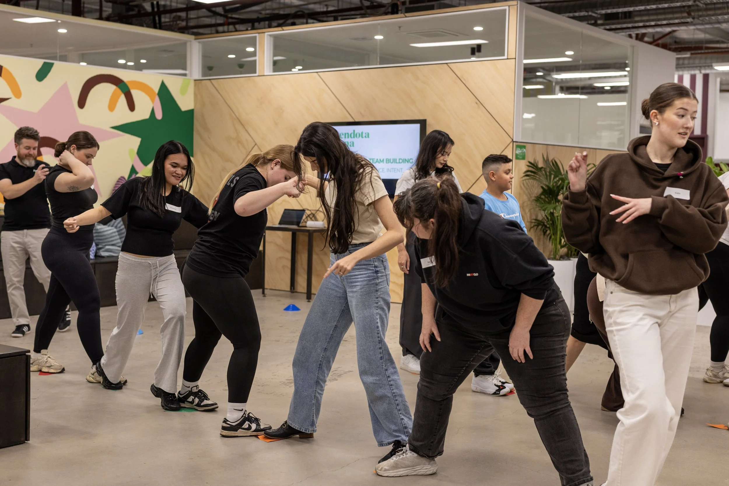 Group of diverse people participating in Field9 team-building exercises in Adelaide, in an indoor space with colourful wall art and digital screens.