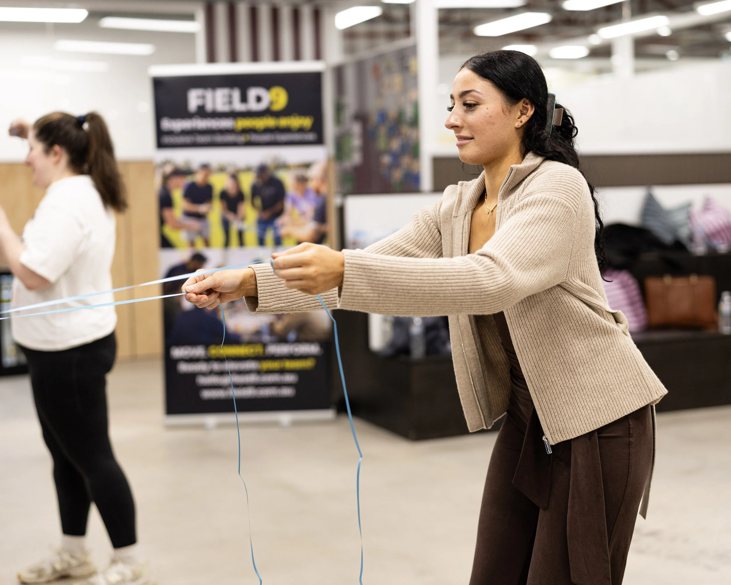A woman with black hair, tan jacket, and earrings is tying blue strings, with others in the background at an indoor Field9 team building event in Adelaide.