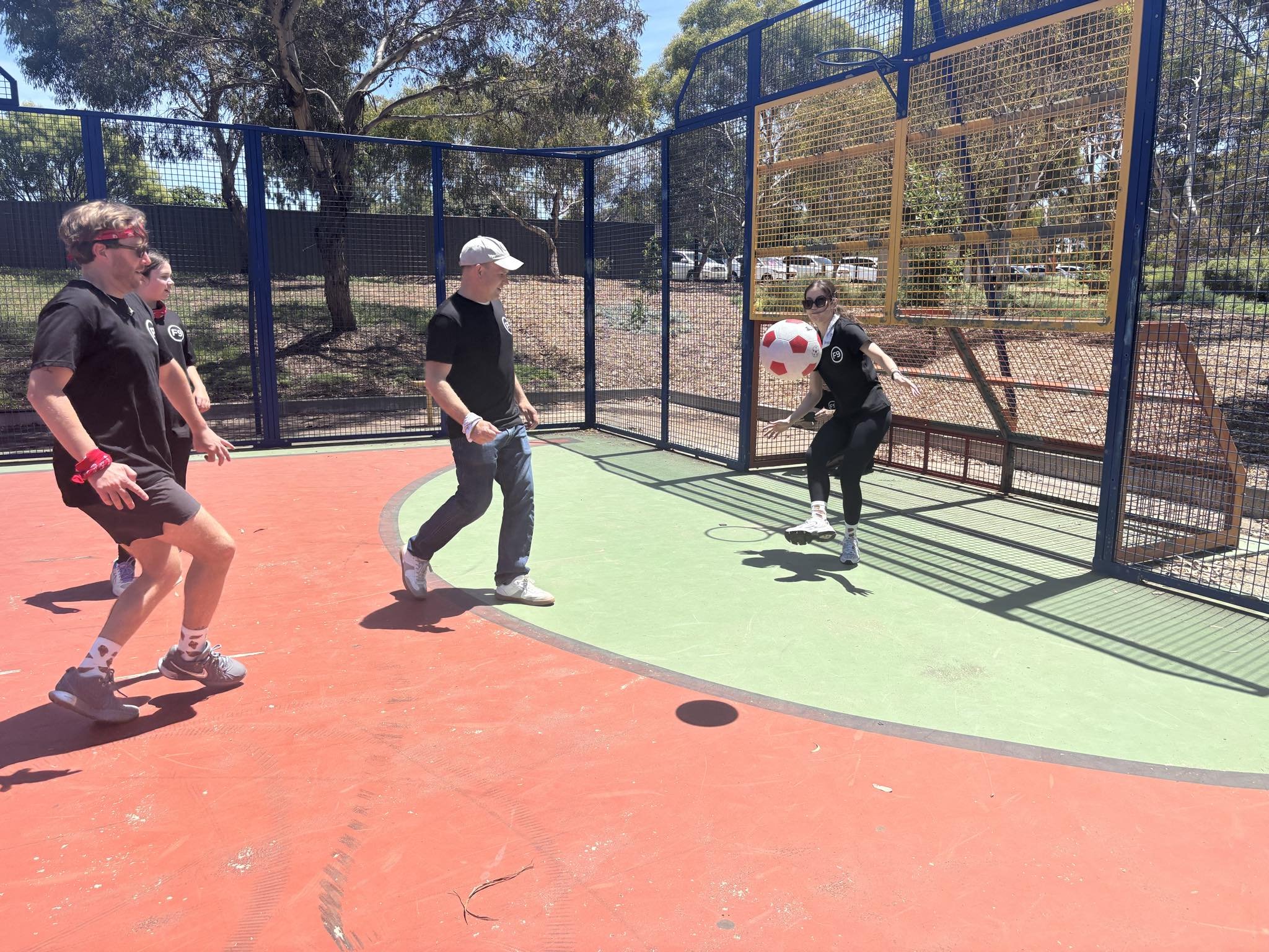 Four people playing soccer on an outdoor court with a female goalie jumping to block a ball, while three others wait for a shot, with trees and parked cars in the background.