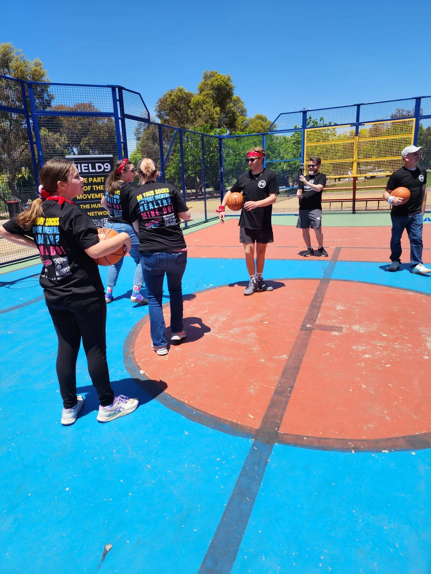 A group of people on an outdoor basketball court, some holding basketballs, with a colorful fence and trees in the background under a bright blue sky, participating in a Field9 Team Building experience in Adelaide.