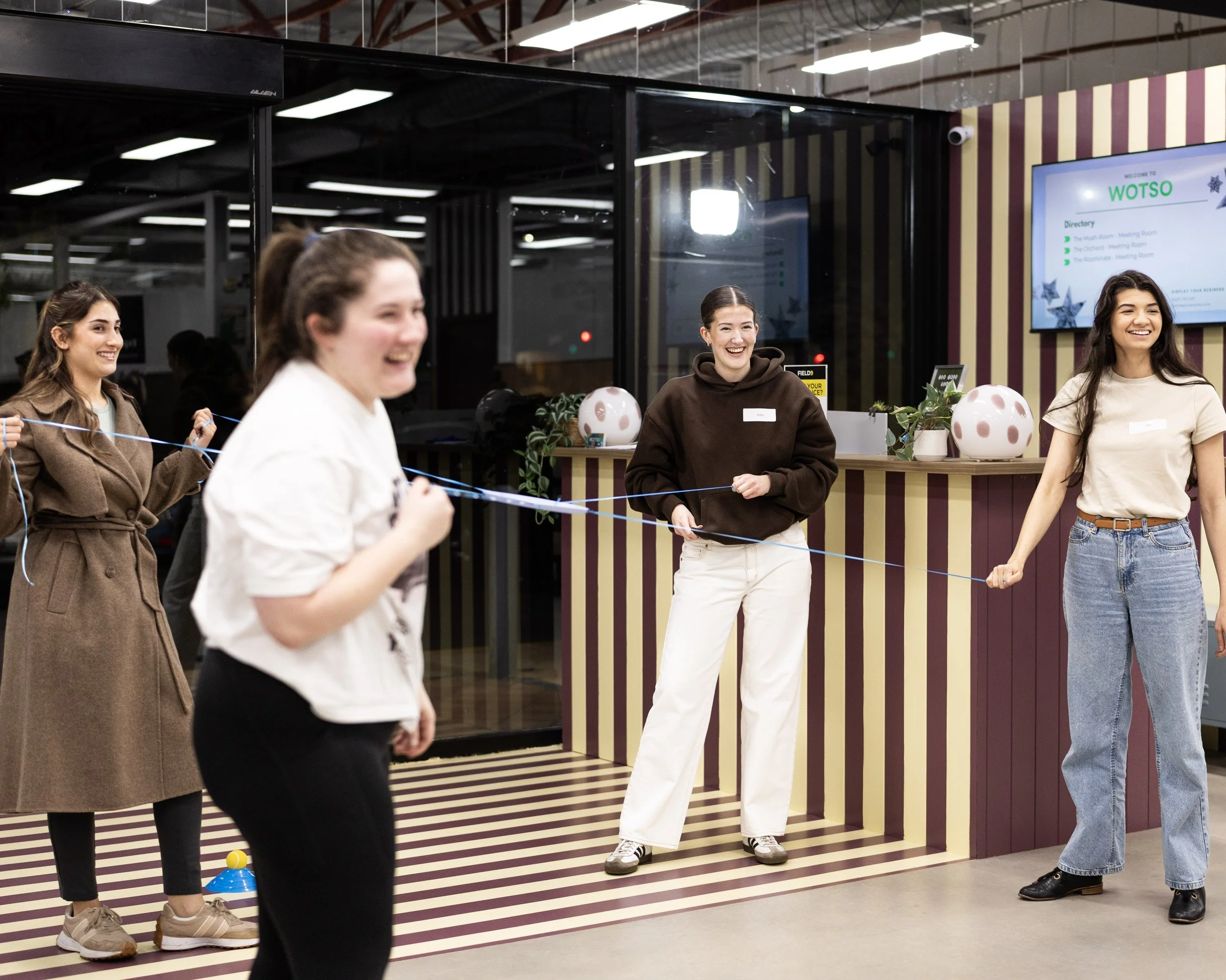Group of people chatting and laughing at an indoor Field9 team building session, with some holding a string of balloons, in a modern space with striped wall and digital directory.