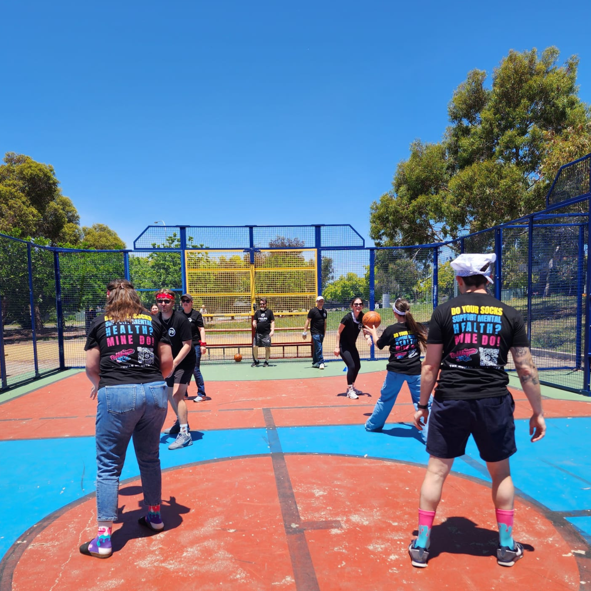 People gathered on an outdoor basketball court with a red surface, surrounded by a blue fence, with trees and a clear blue sky in the background. The individuals are participating in a community event, with some wearing black T-shirts with colorful t