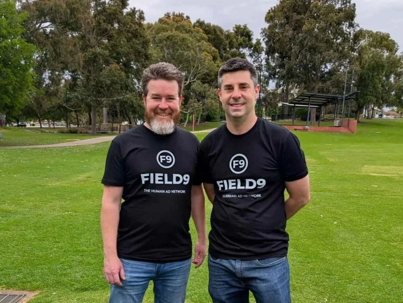 Two men standing outdoors on a grassy field, wearing matching black T-shirts with the text 'FIELD9 THE HUMAN AD NETWORK' and a circle with 'F9' printed inside.