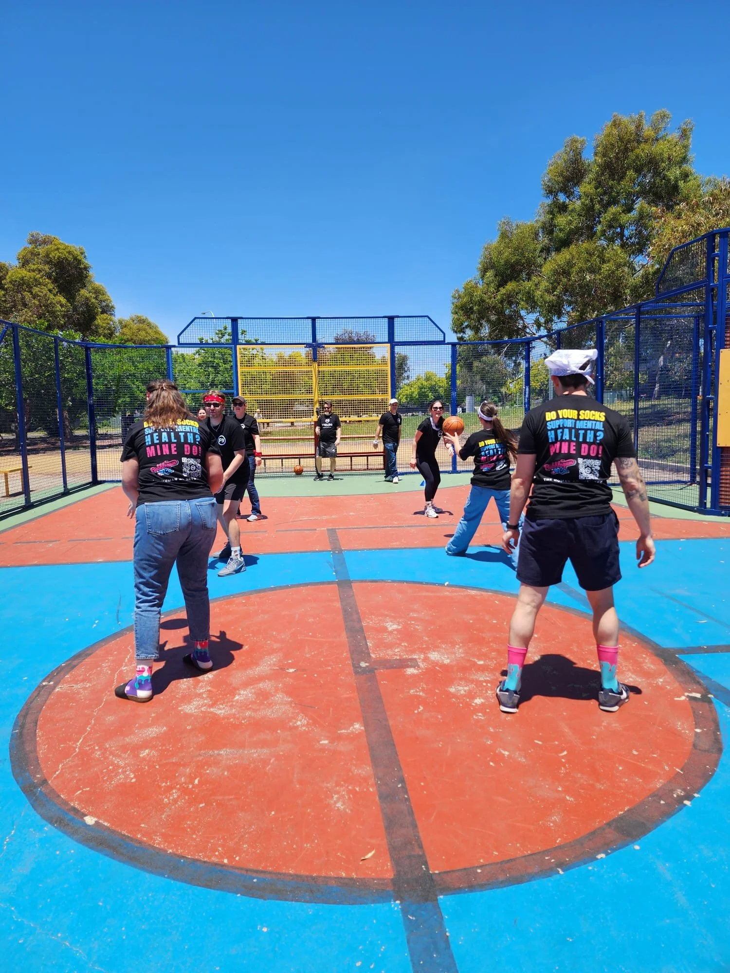 People playing basketball on an outdoor court with colorful markings, surrounded by a tall blue fence, and trees in the background under a clear blue sky, participating in a Field9 Team Building experience in Adelaide.